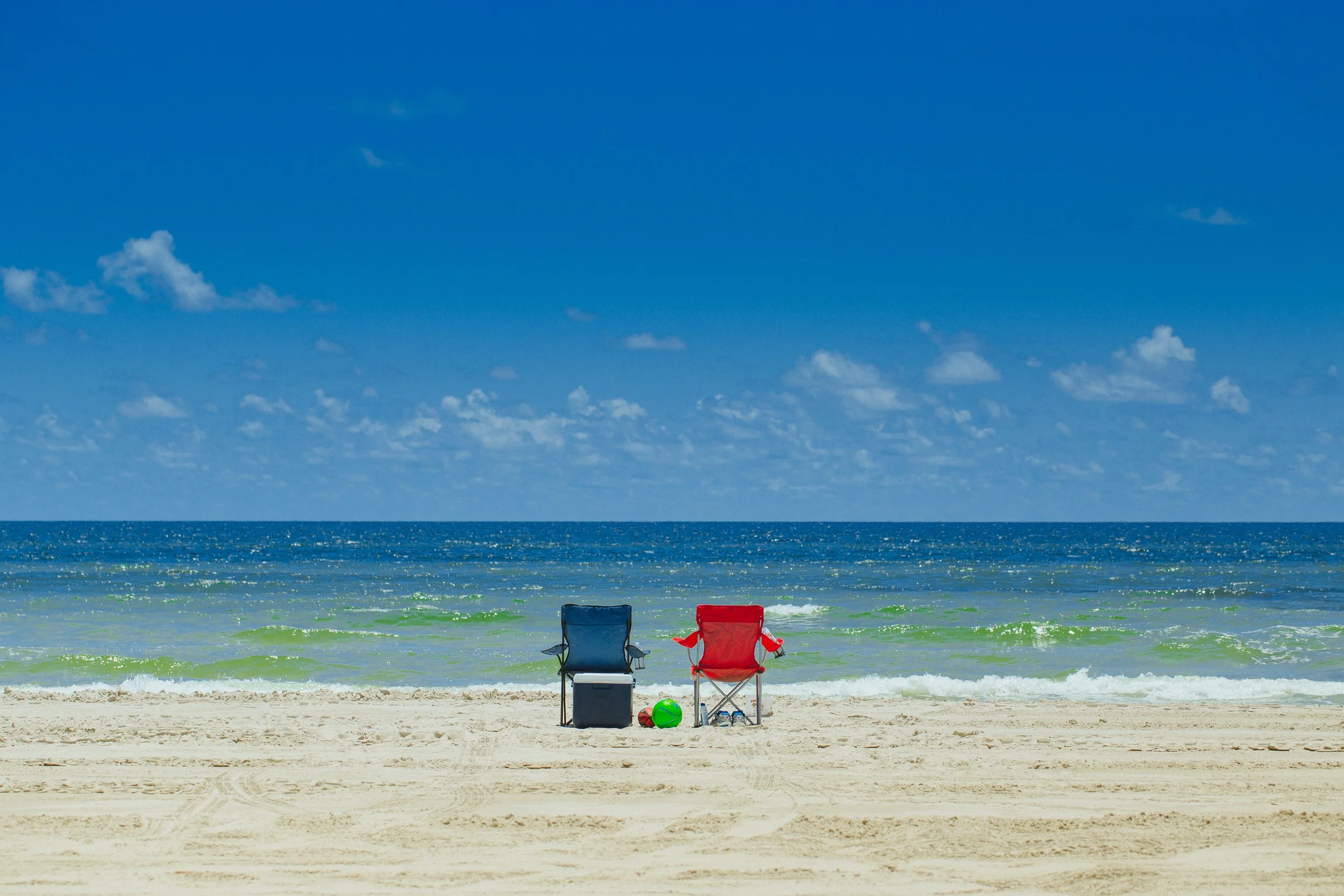 Two beach chairs, one blue and one red, sit on the sandy beach facing the ocean, with a green ball and a red ball on the sand in front of the chairs. The ocean waves and a blue sky with scattered clouds are in the background.