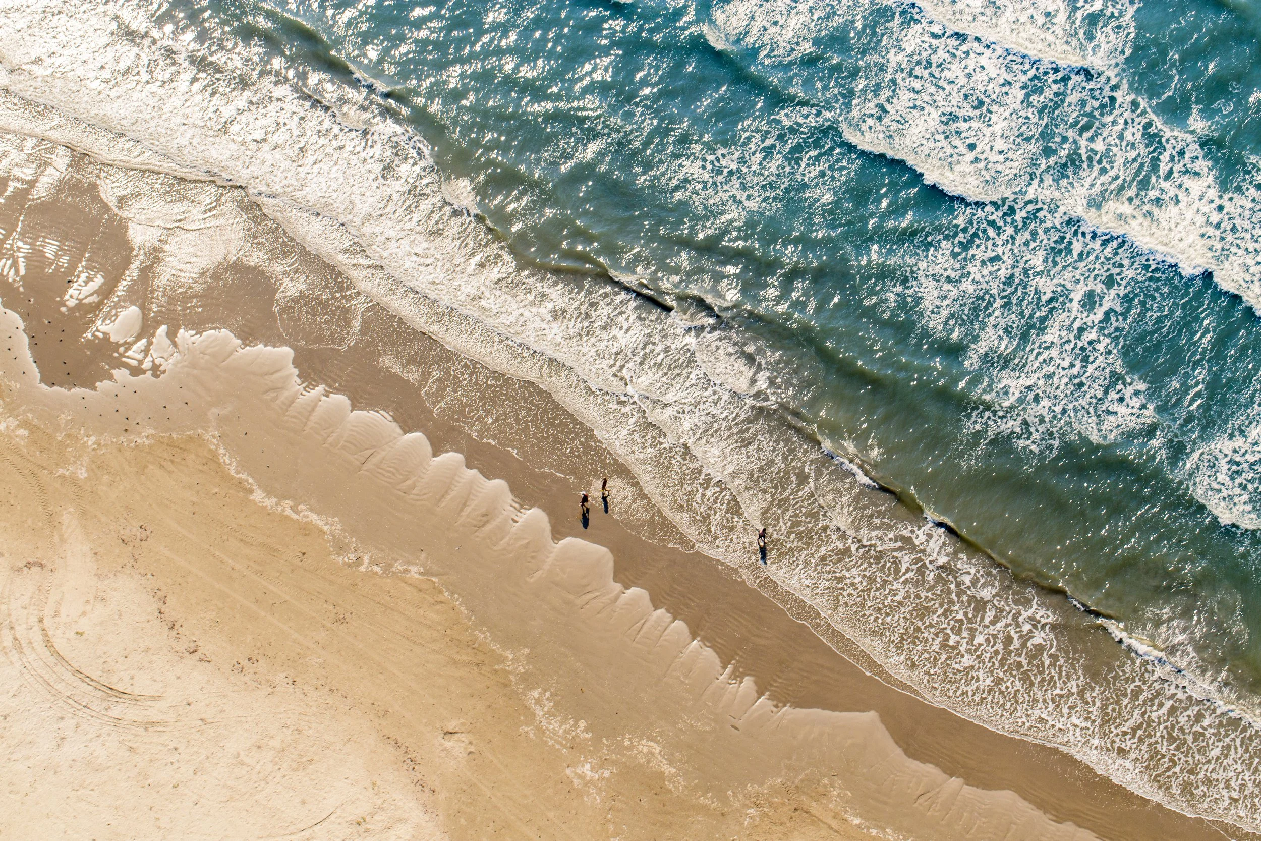 An aerial view of a sandy beach with waves crashing onto the shore and three people walking along the water's edge.