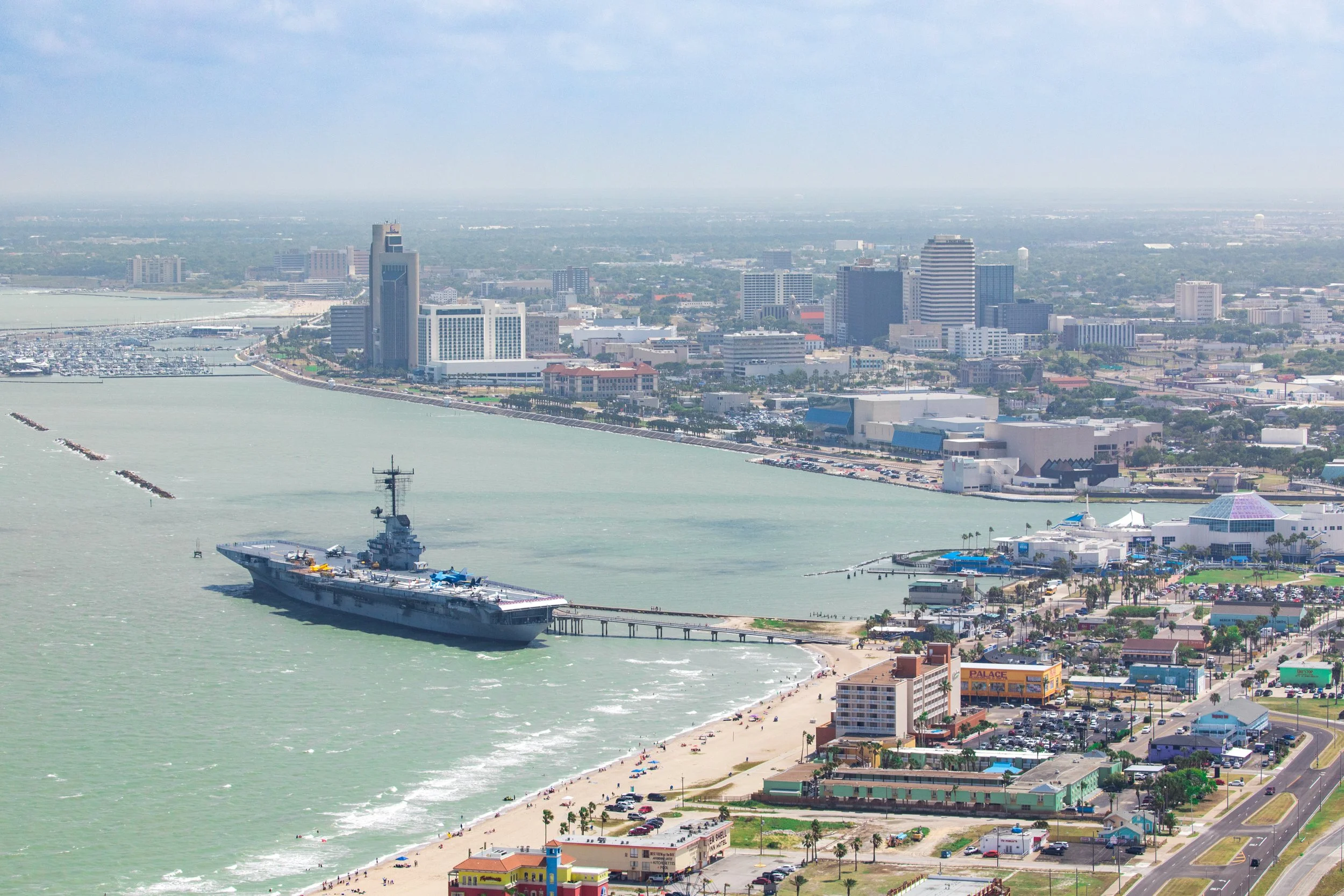 Aerial view of a city skyline near the coast with a large naval aircraft carrier and downtown buildings, near a beach.