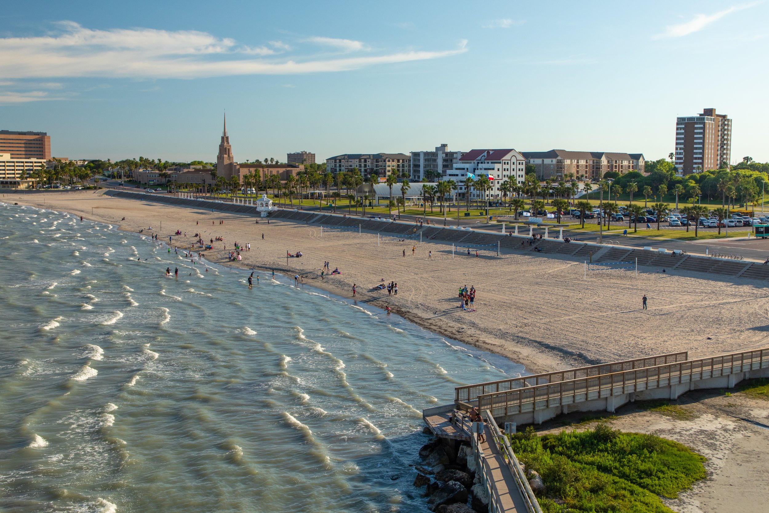 A beach with people enjoying the water and sand, with a pier and a cityscape in the background, including tall buildings and a church with a steeple, during daytime.