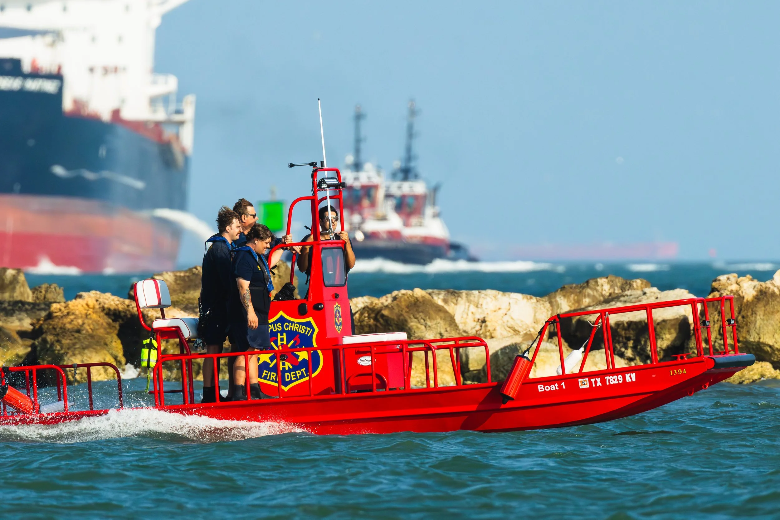 Firefighters on a red rescue boat near rocky shoreline with large ships in the background