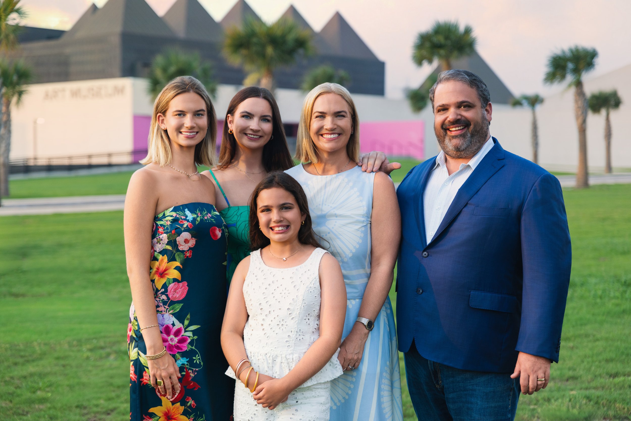 Family of five, including two young women, a young girl, a woman, and a man, standing outdoors on a grassy area with palm trees and modern building in the background, smiling at the camera.
