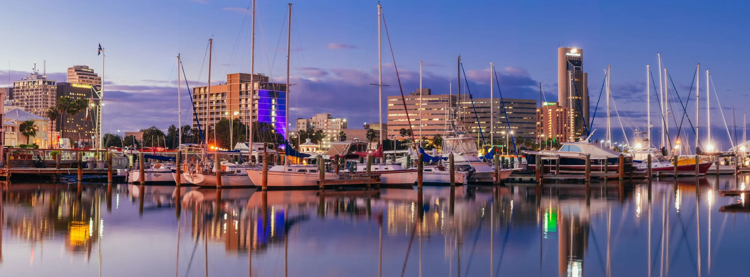 View of a marina with sailboats and yachts docked, city buildings illuminated in the background, and a calm water reflection during dusk.