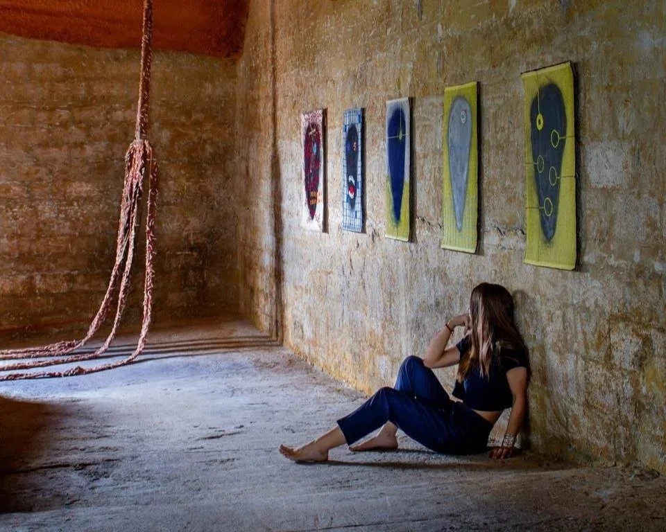A young woman with long hair, dressed in a black shirt and blue pants, sits on the floor in a rustic room with textured yellowish walls. She leans back against the wall and looks at a series of colorful, abstract paintings hanging on the wall.