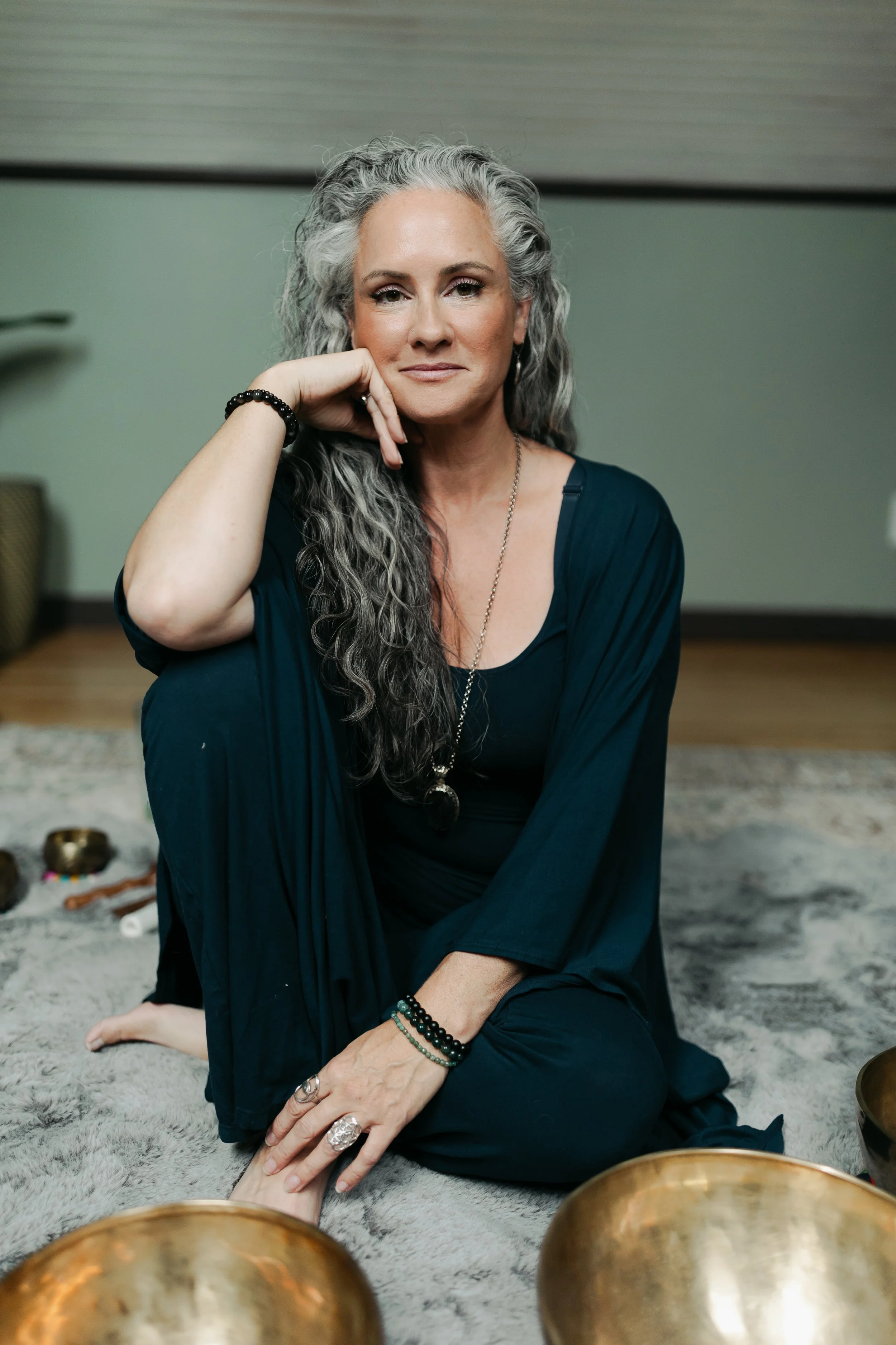 A woman with long gray hair, wearing a dark blue dress and jewelry, sits on a soft gray rug in a room, looking directly at the camera with a calm expression, resting her head on her hand, surrounded by sound bowls, indicating a peaceful setting.