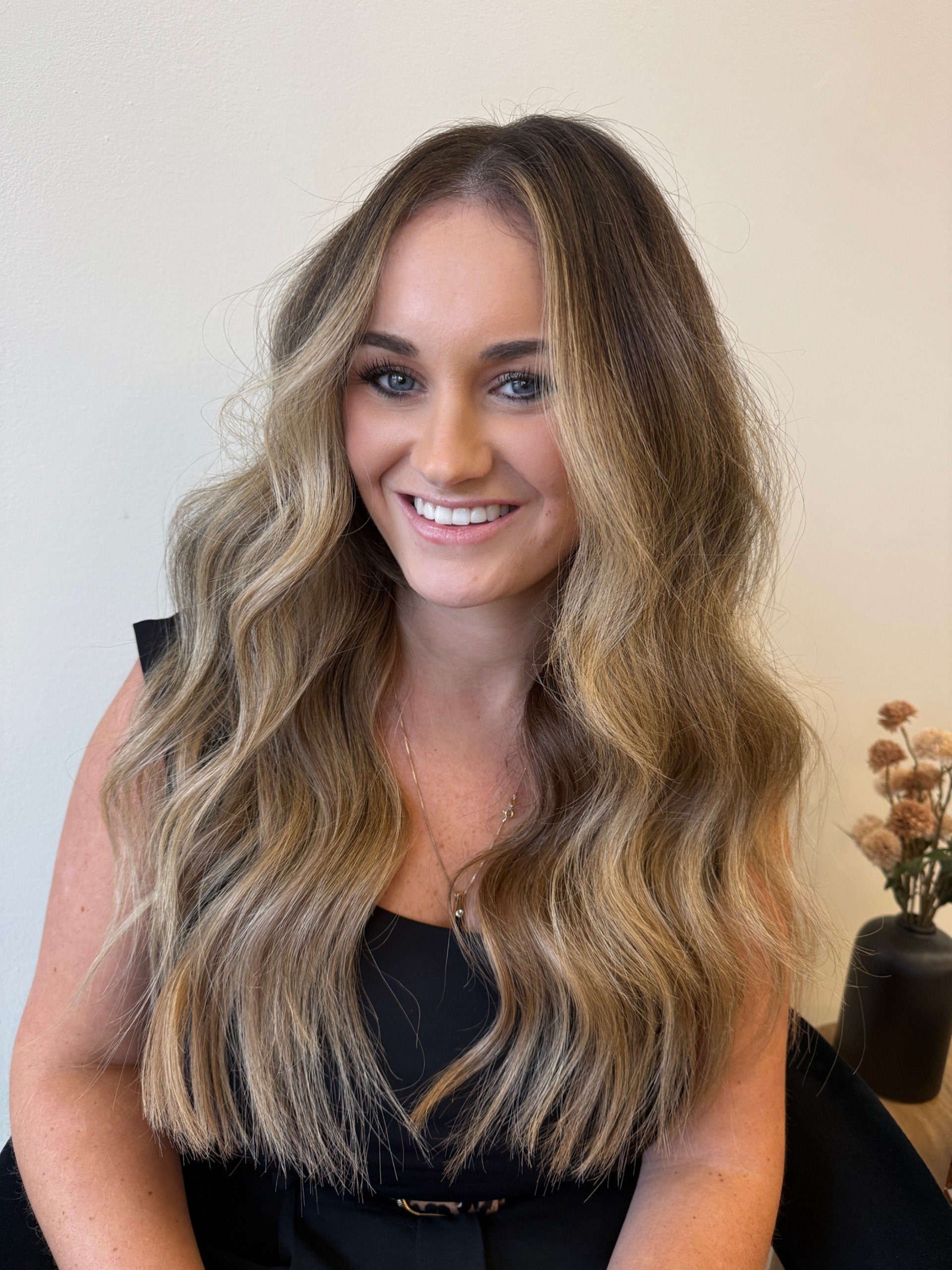 A woman with long, wavy, light brown hair with blonde highlights, smiling and wearing a black top.