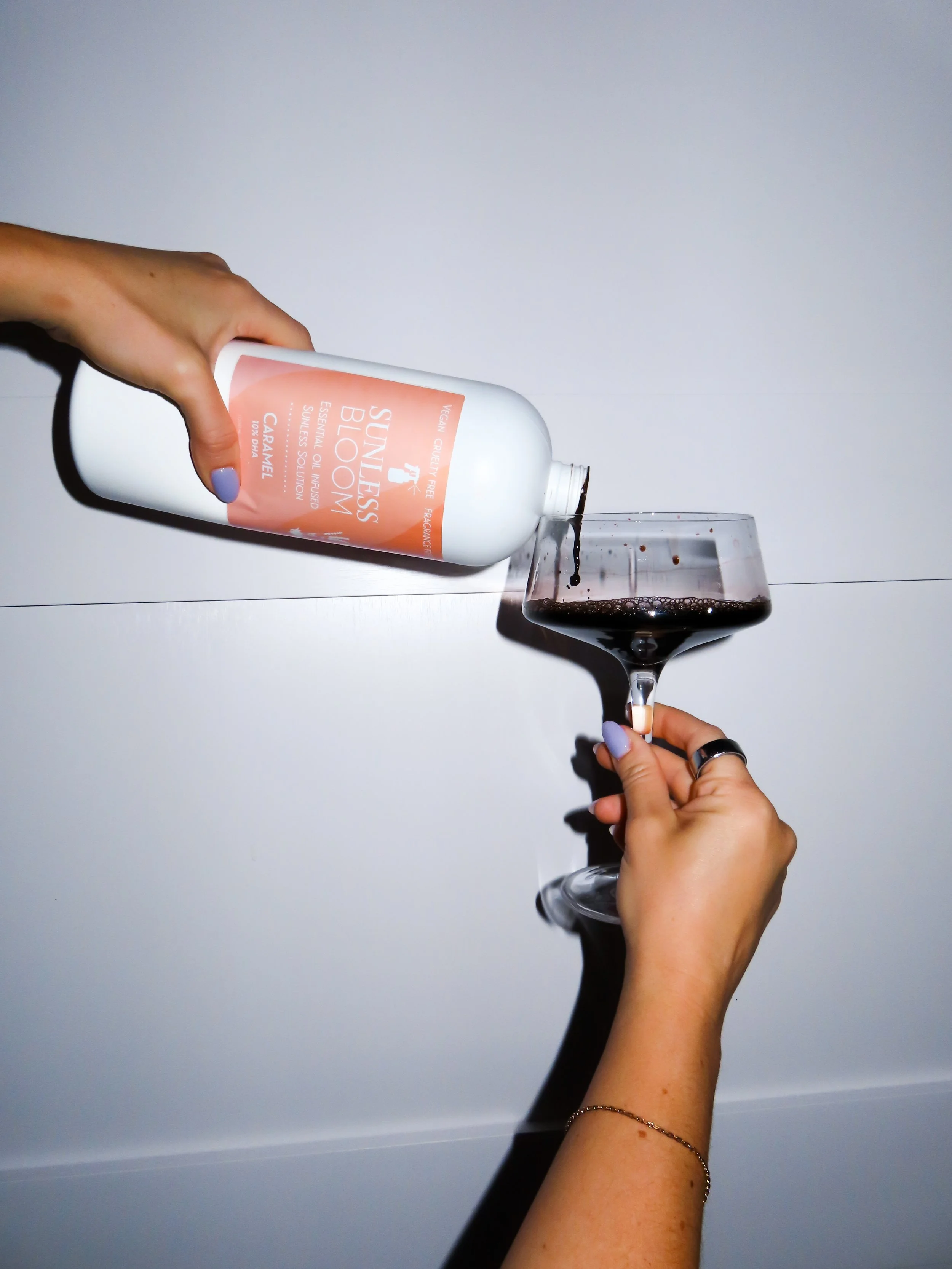 A person with light skin and painted nails is pouring black liquid from a white bottle labeled 'Sunless Bloom Caramel' into a glass of dark liquid, possibly wine, against a plain light background.