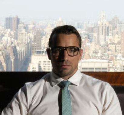 Man wearing white shirt, green tie, and glasses sitting in front of a city skyline background.
