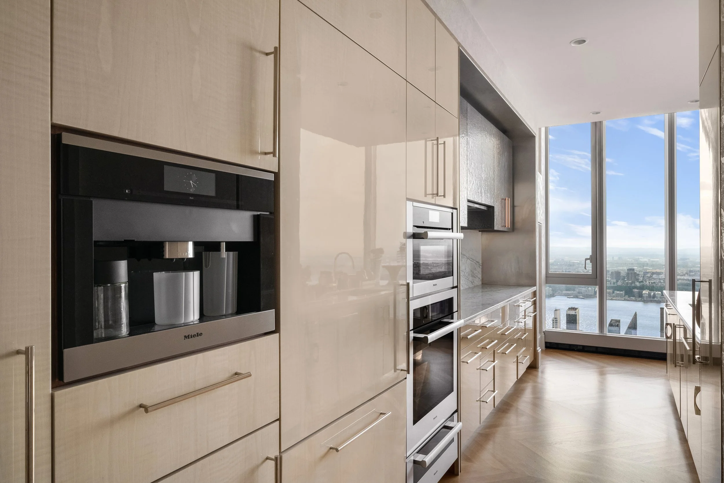 Modern kitchen with built-in appliances and large windows showing a cityscape and blue sky.