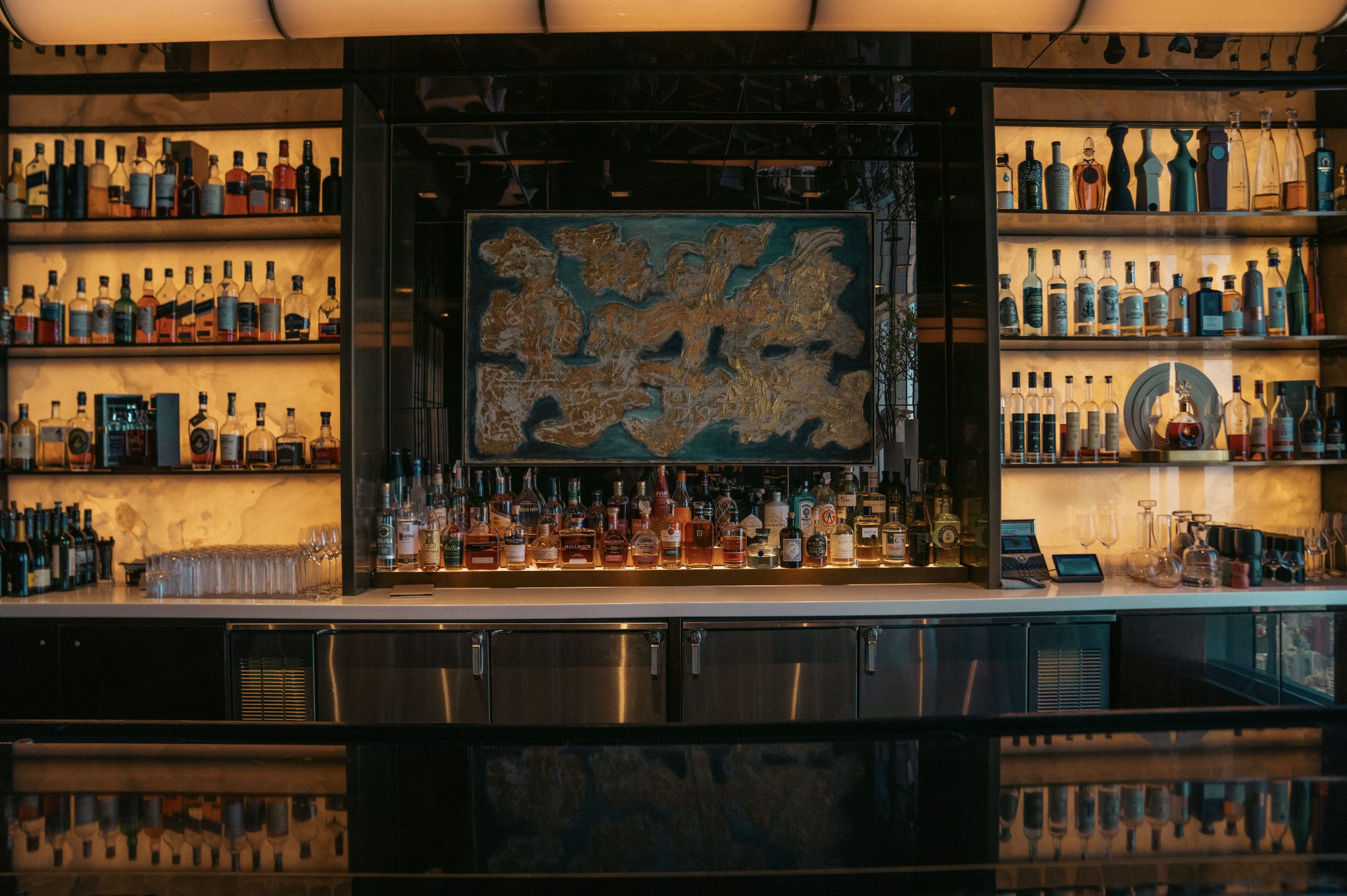 Bar counter with shelves of liquor bottles lit by warm lighting in a modern bar.