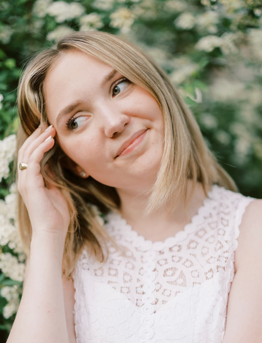 A young woman with blonde hair touching her head, smiling gently, in front of green foliage and white flowers, wearing a white lace top.