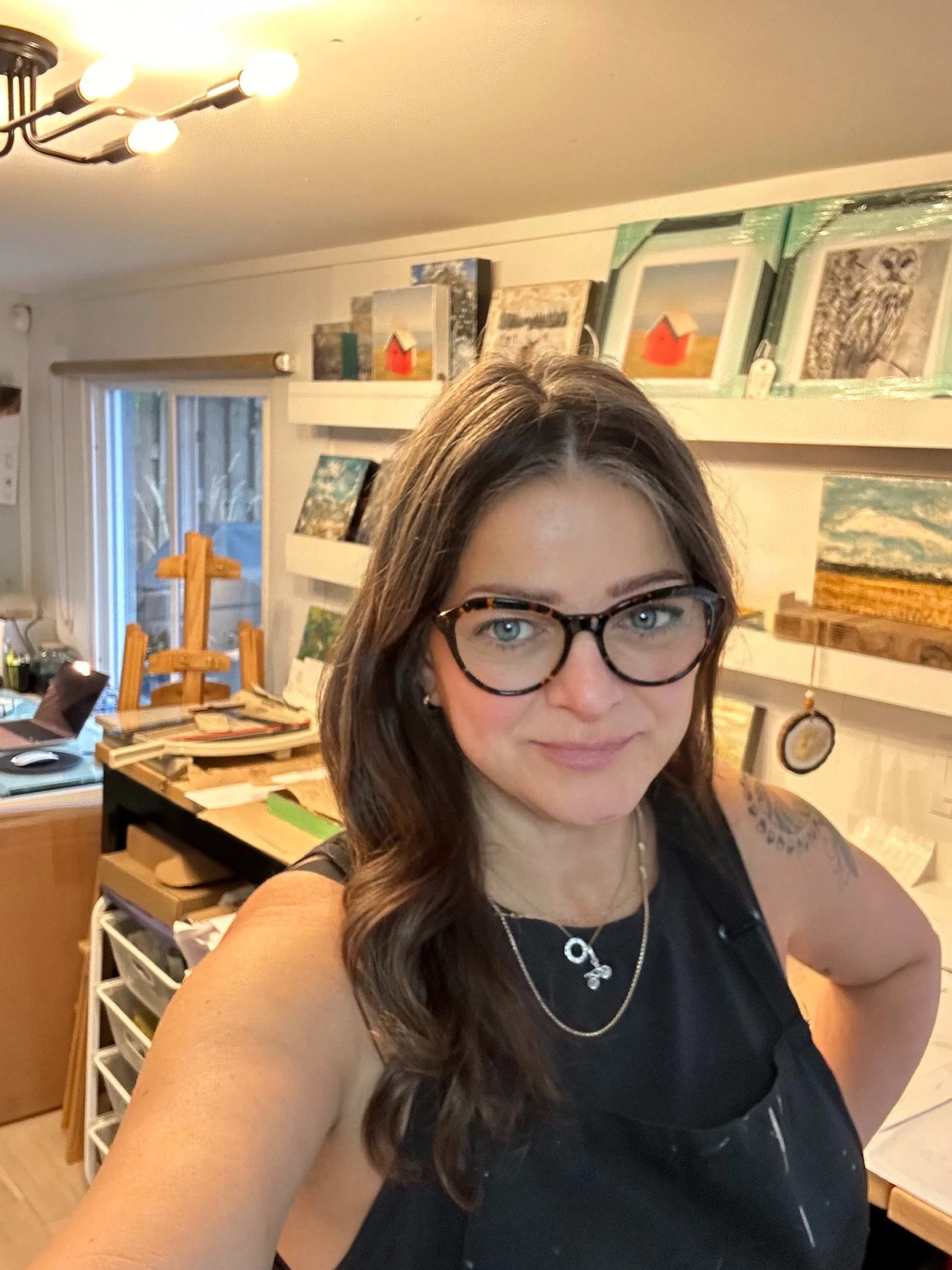 An artist in her art studio filled with paintings, a bookshelf, and various art supplies.