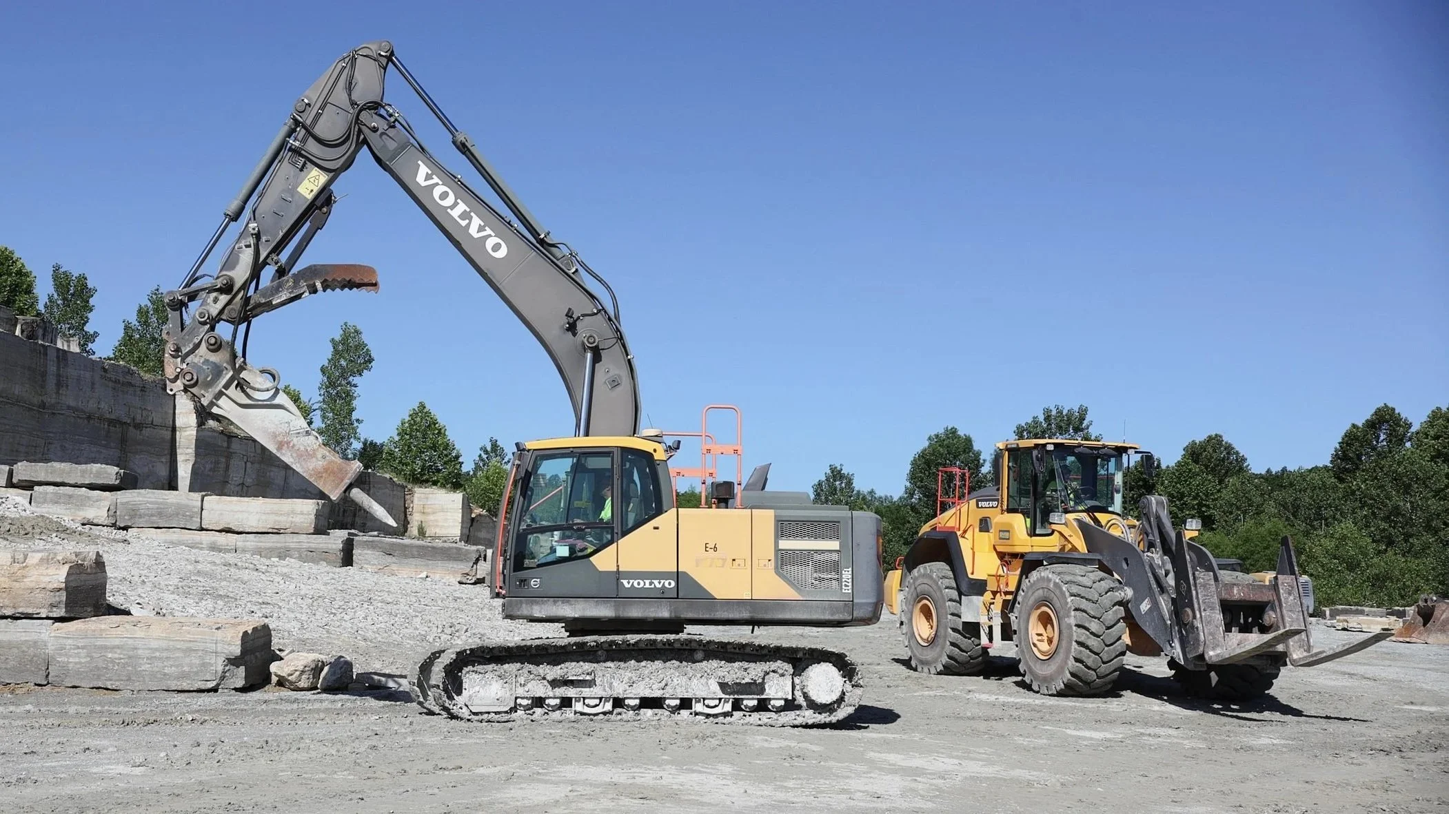A construction site with a large excavator and a front loader, working on a stone or concrete structure, under a clear blue sky.