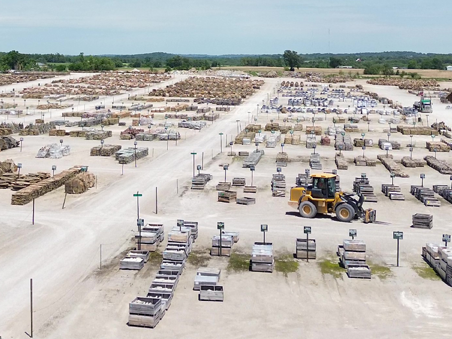 A large outdoor storage yard with stacks of stone slabs and blocks, a yellow forklift moving pallets, and cars parked in the background under a cloudy sky.
