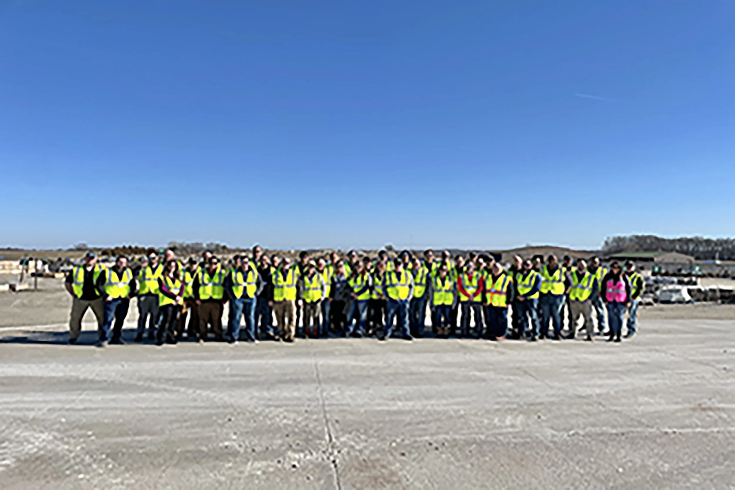 A large group of people wearing yellow safety vests standing outdoors in a line on a sunny day with a clear blue sky.