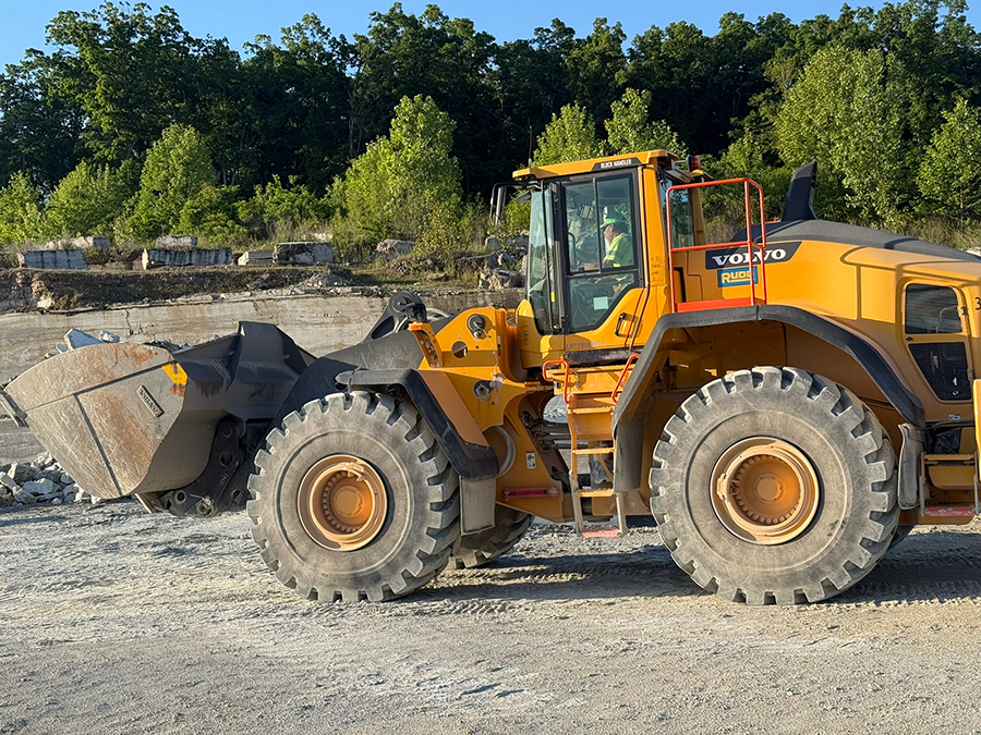A yellow Volvo construction vehicle, likely a wheel loader, on a dirt construction site with a backdrop of trees.