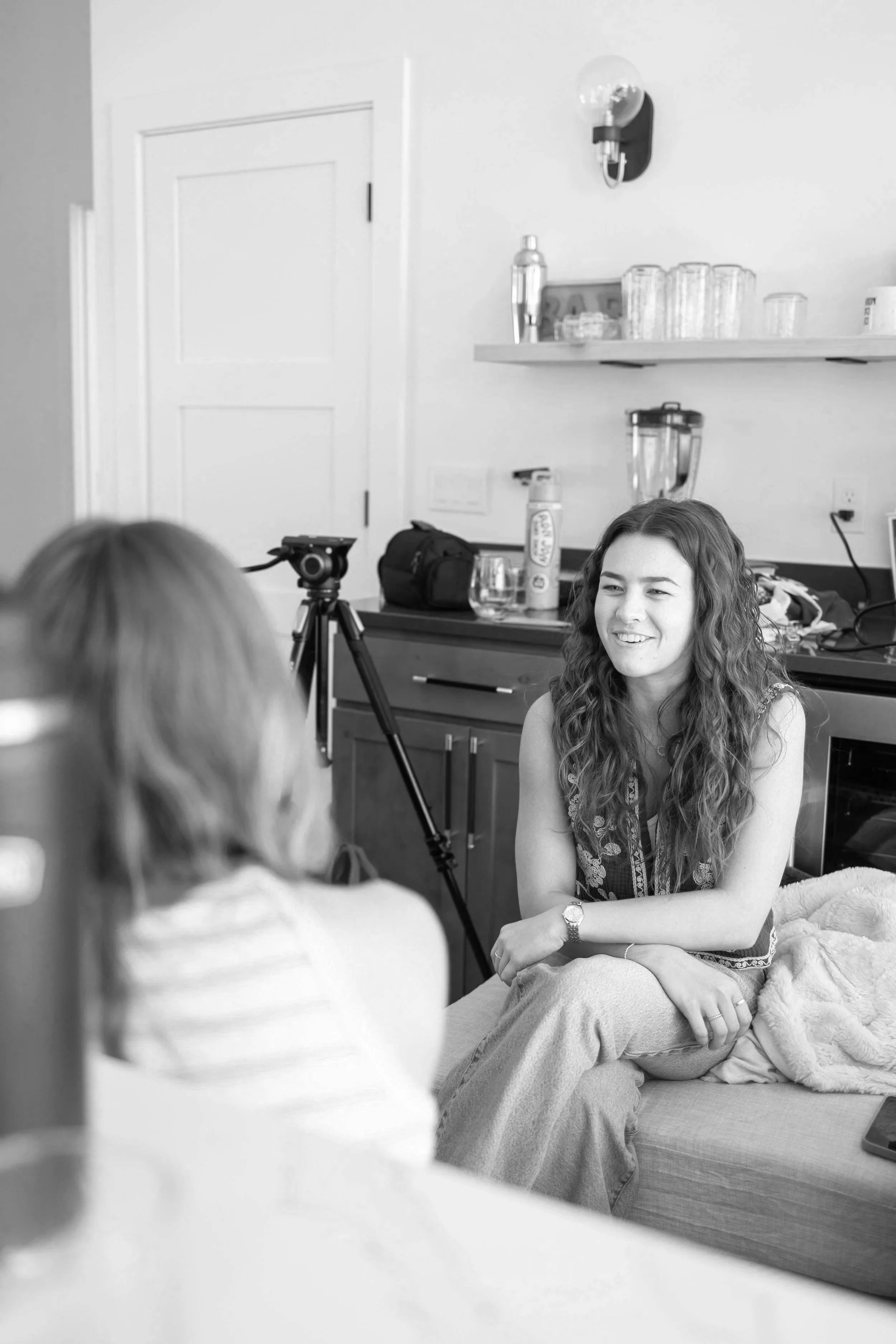 A woman with long, wavy hair smiling while sitting on a bed or sofa, having a conversation with children in a room with shelves, glasses, a blender, and a camera on a tripod in the background.