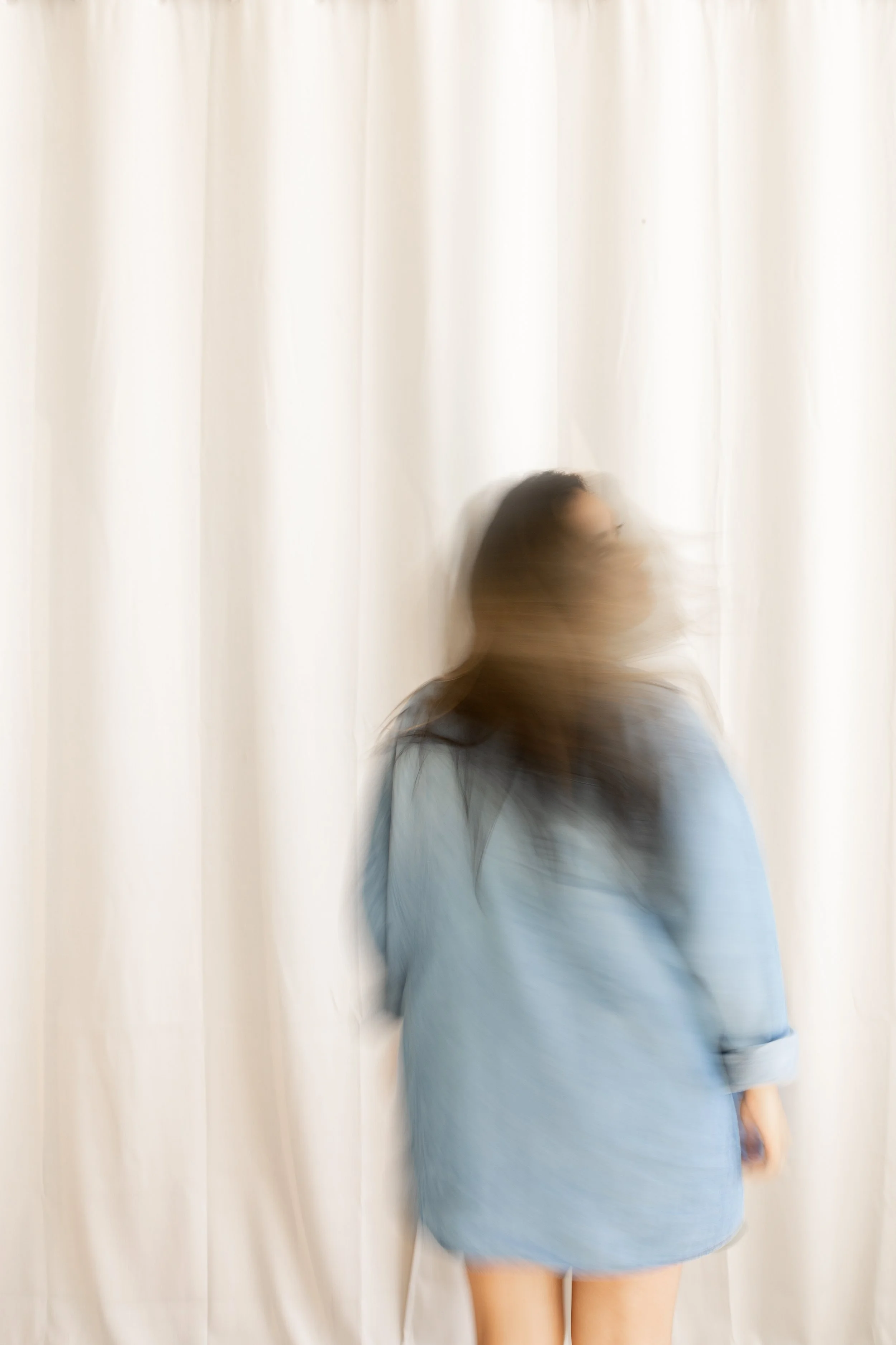 A person stands in front of a white curtain wearing a light blue dress, with motion blur affecting the person's face and body.