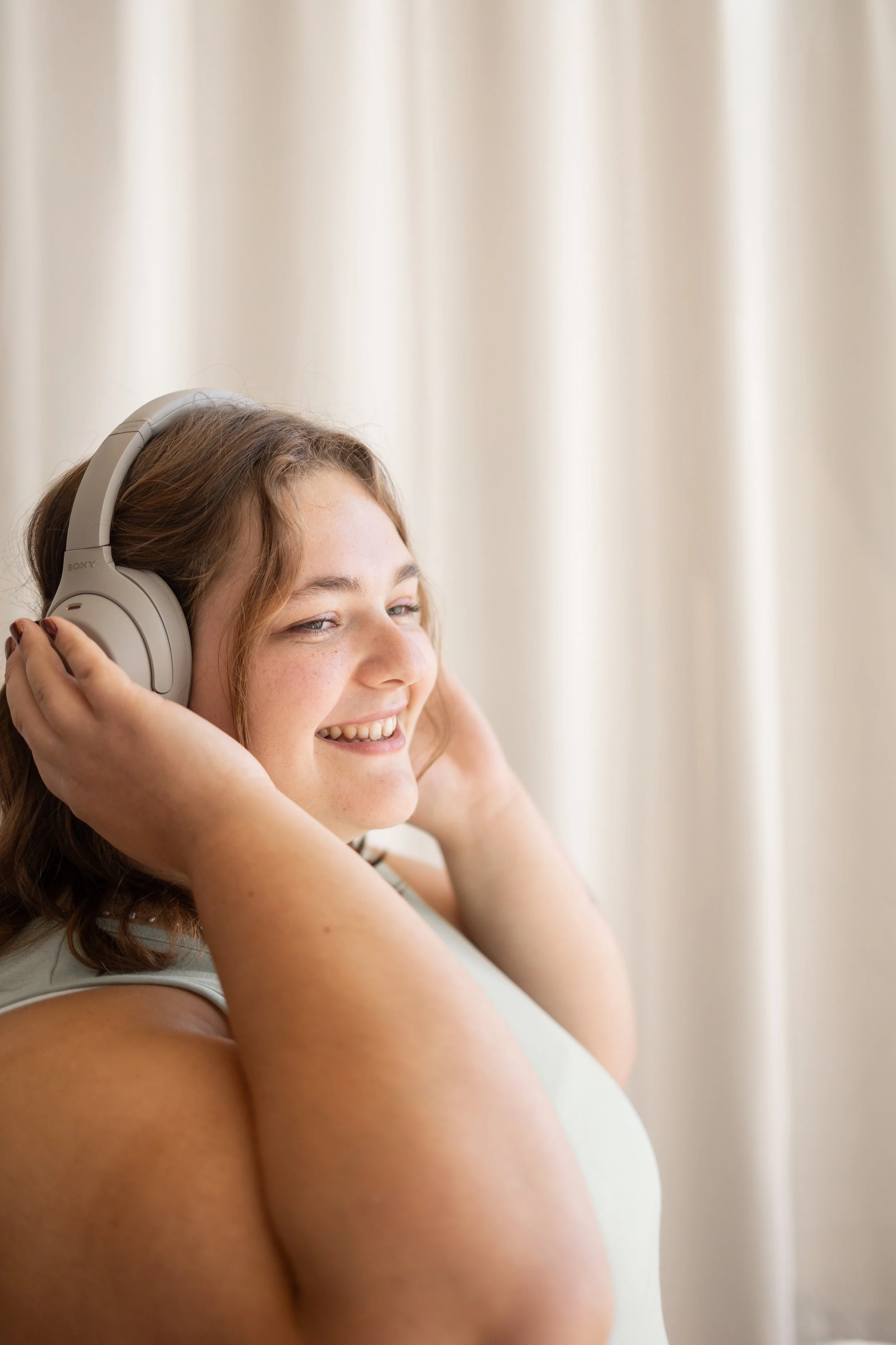 A young woman with brunette hair smiling while listening to music with over-ear headphones, sitting in front of a cream-colored curtain.