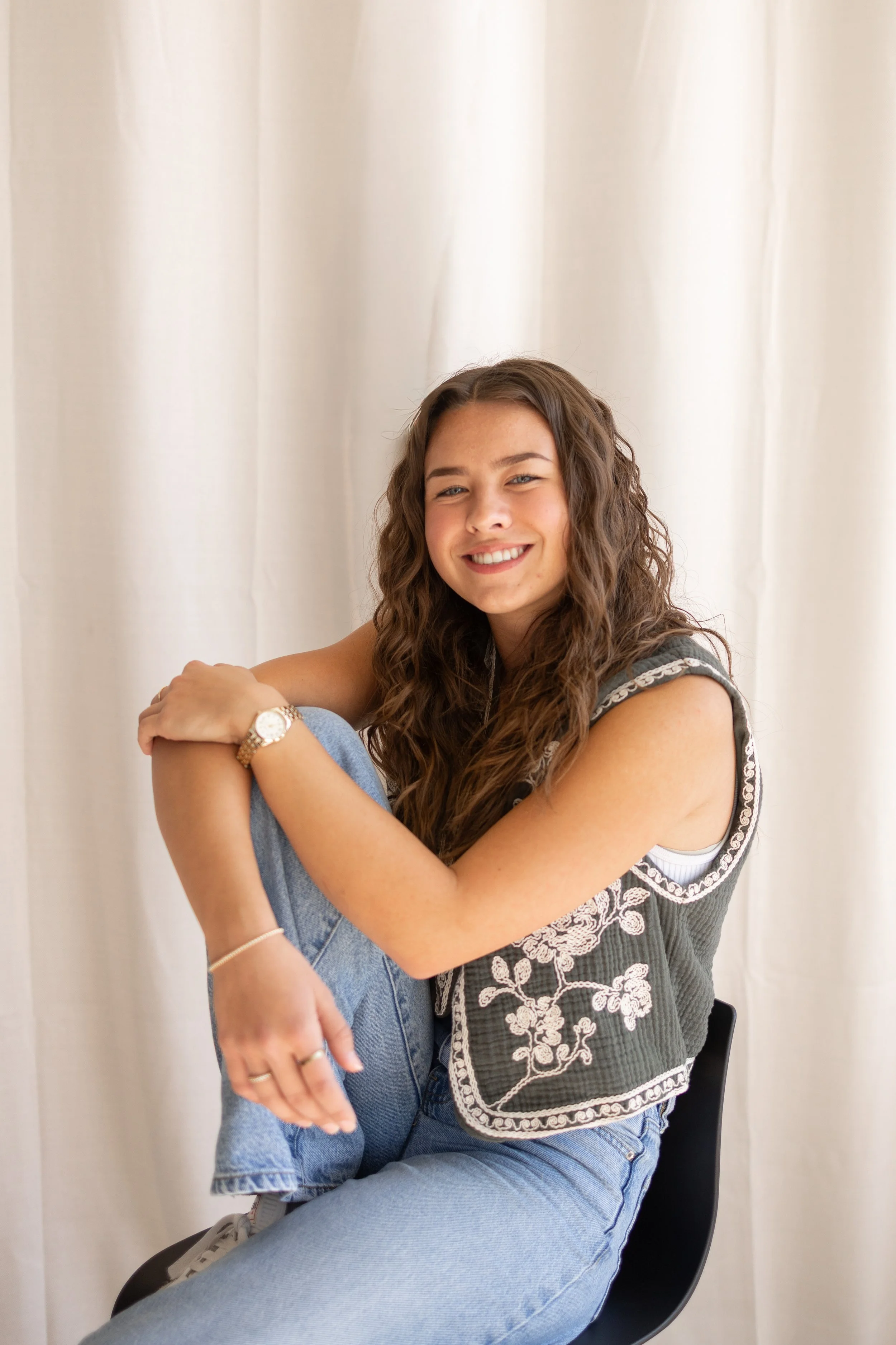A young woman with long, wavy brown hair is sitting on a black chair in front of a plain, off-white curtain. She is smiling at the camera and wearing a grey sleeveless top with embroidered white floral patterns and light blue jeans. She has a gold wristwatch on her left wrist and a gold bracelet on her right wrist.
