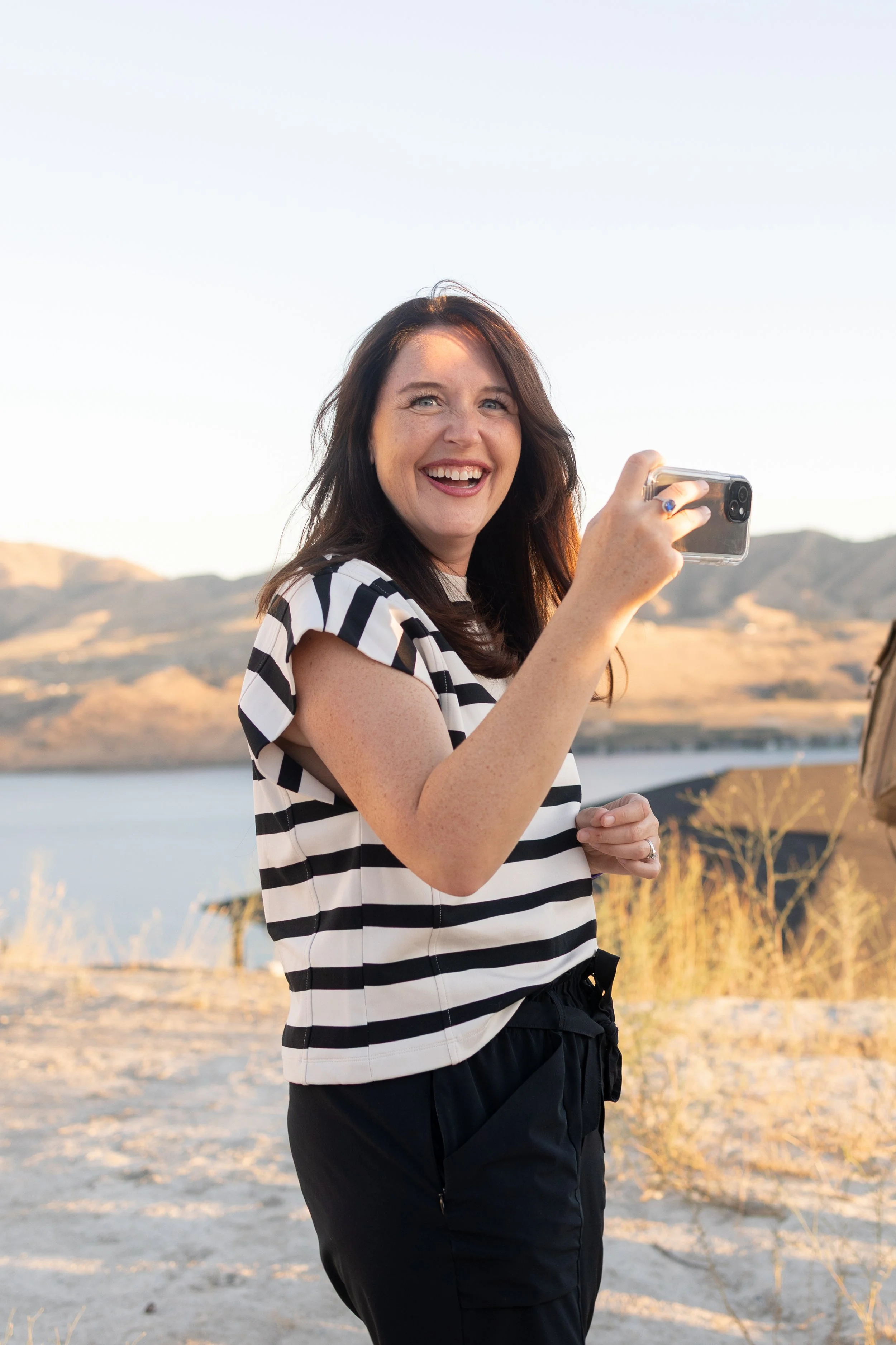 A woman with dark hair and blue eyes smiling while holding a smartphone outdoors, with mountains and water in the background during sunset.