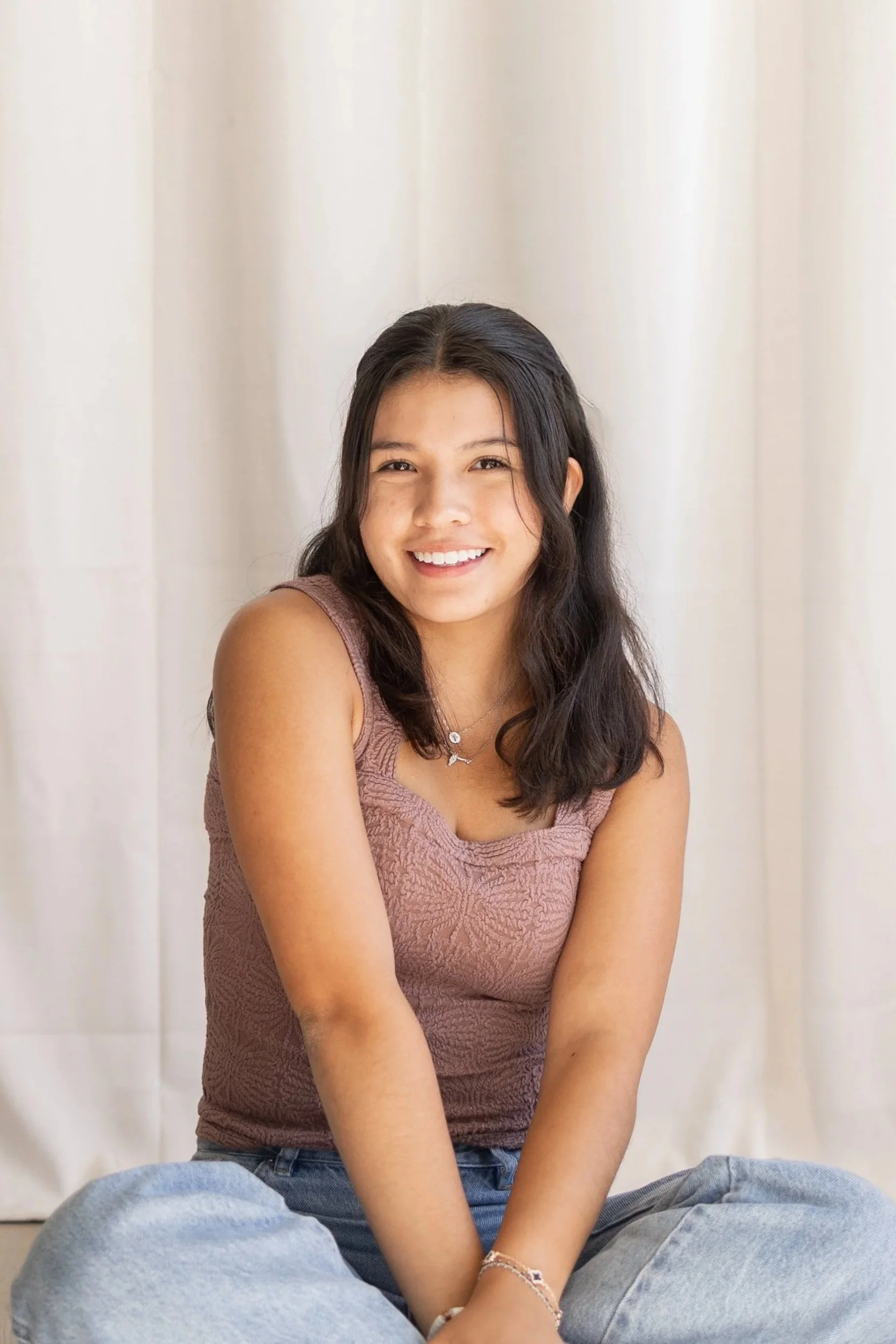 Young woman with long dark hair, smiling, sitting cross-legged against a light background, wearing a sleeveless textured top and jeans.