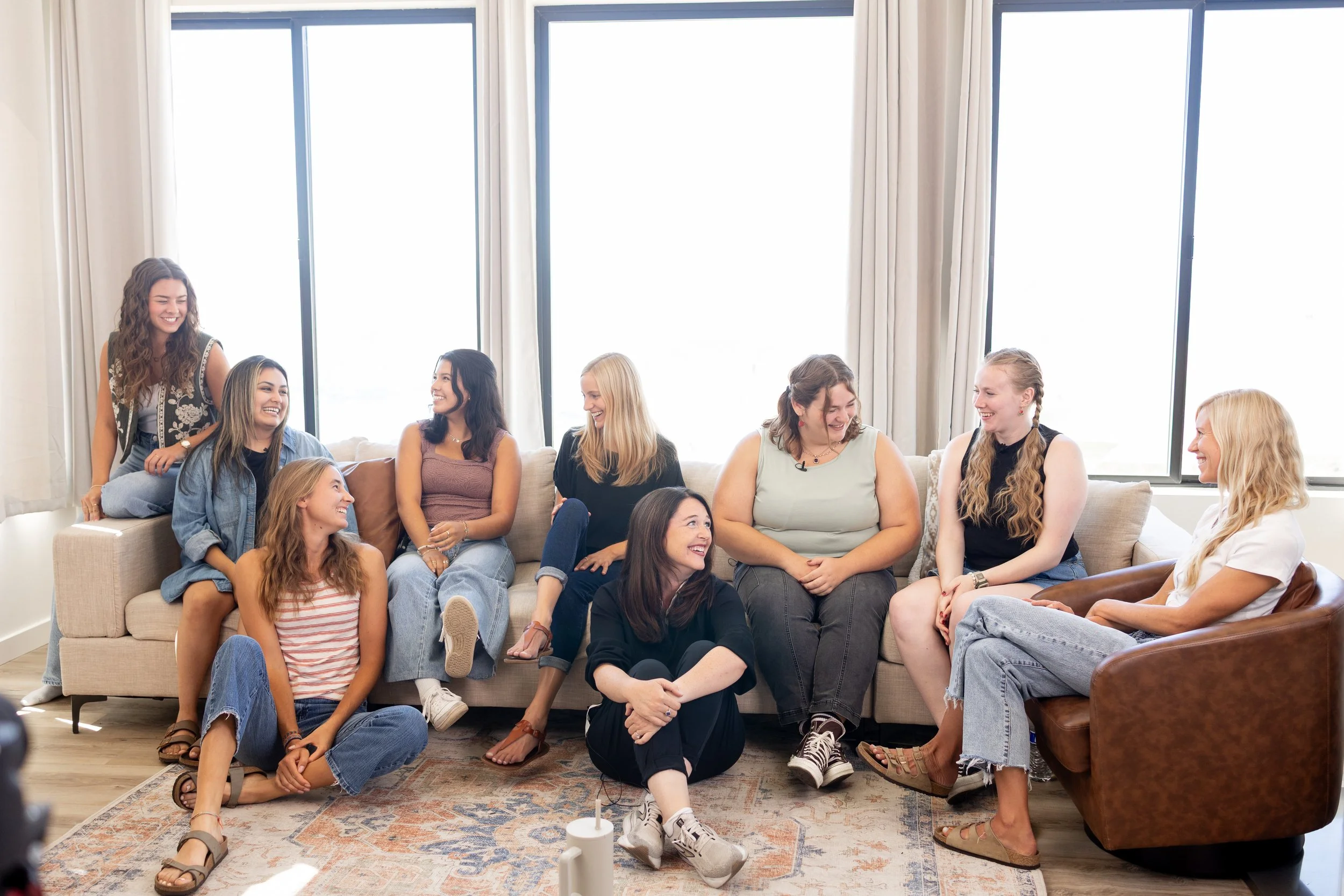 Group of women sitting and talking together on a sofa in a bright living room with large windows.