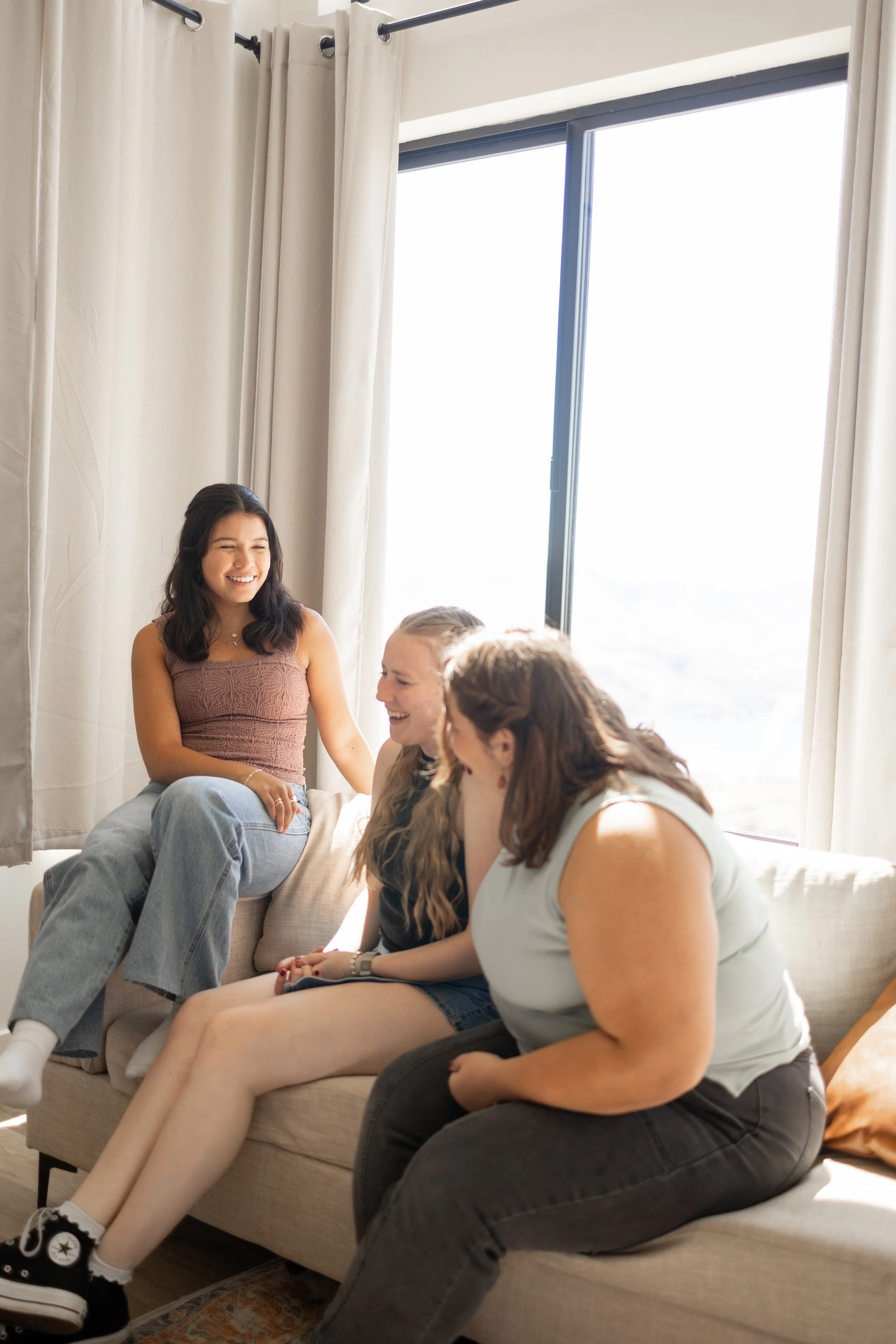 A group of young women socializing and smiling in a bright living room with large windows and white curtains. One woman sits on a sofa, others sit or lean on the sofa, and they are engaged in conversation.