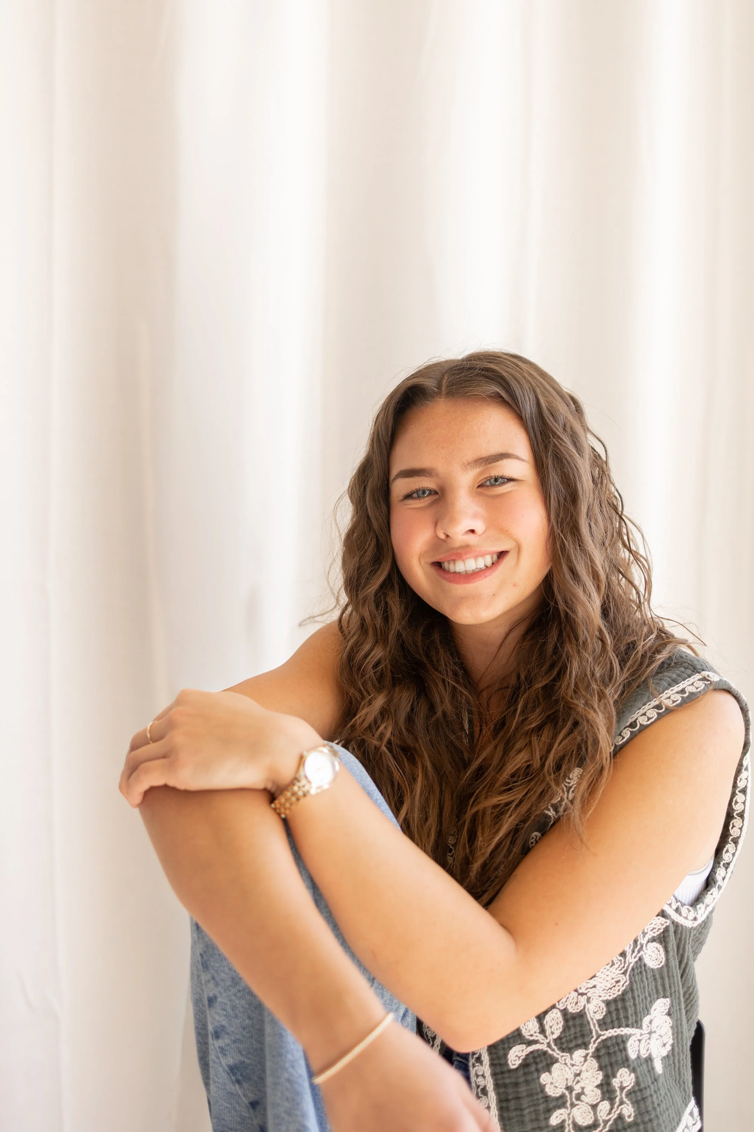 A young woman with long, curly brown hair and blue eyes smiling at the camera, sitting near a cream-colored curtain, wearing a grey embroidered top, a wristwatch, and a bracelet.