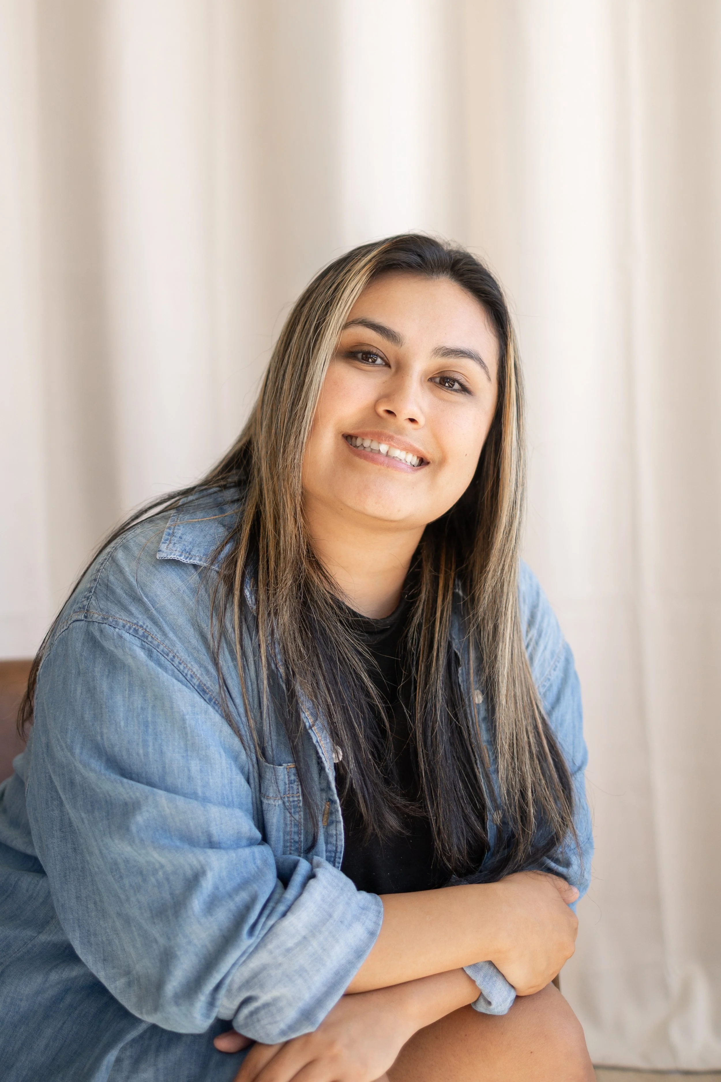 A woman with long hair, wearing a black top and a denim jacket, smiling and sitting with her arms crossed in front of a neutral background.