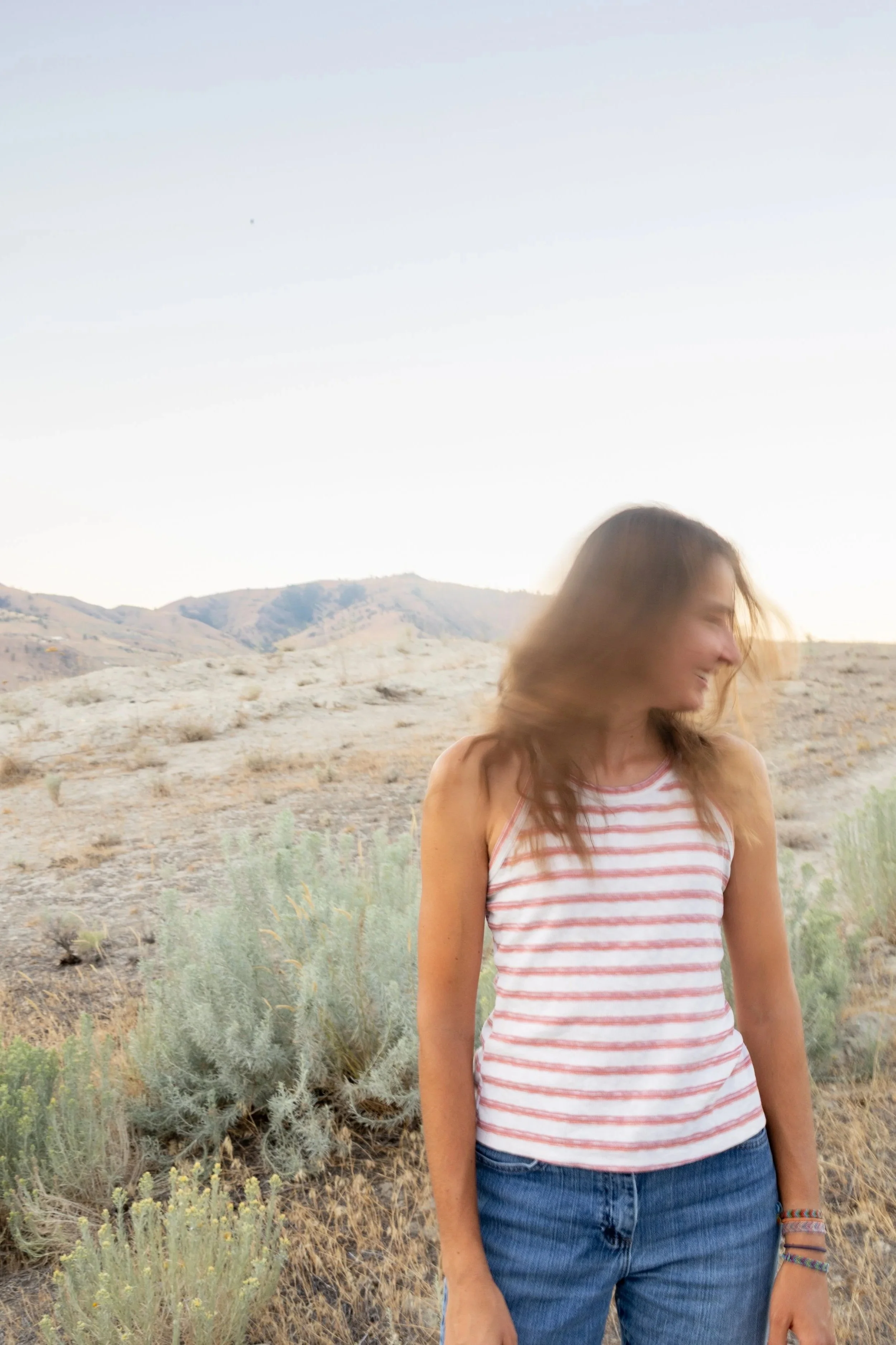 A woman standing in a desert landscape with mountains in the background, wearing a striped sleeveless top and jeans, smiling with her hair blowing in the wind during the daytime.