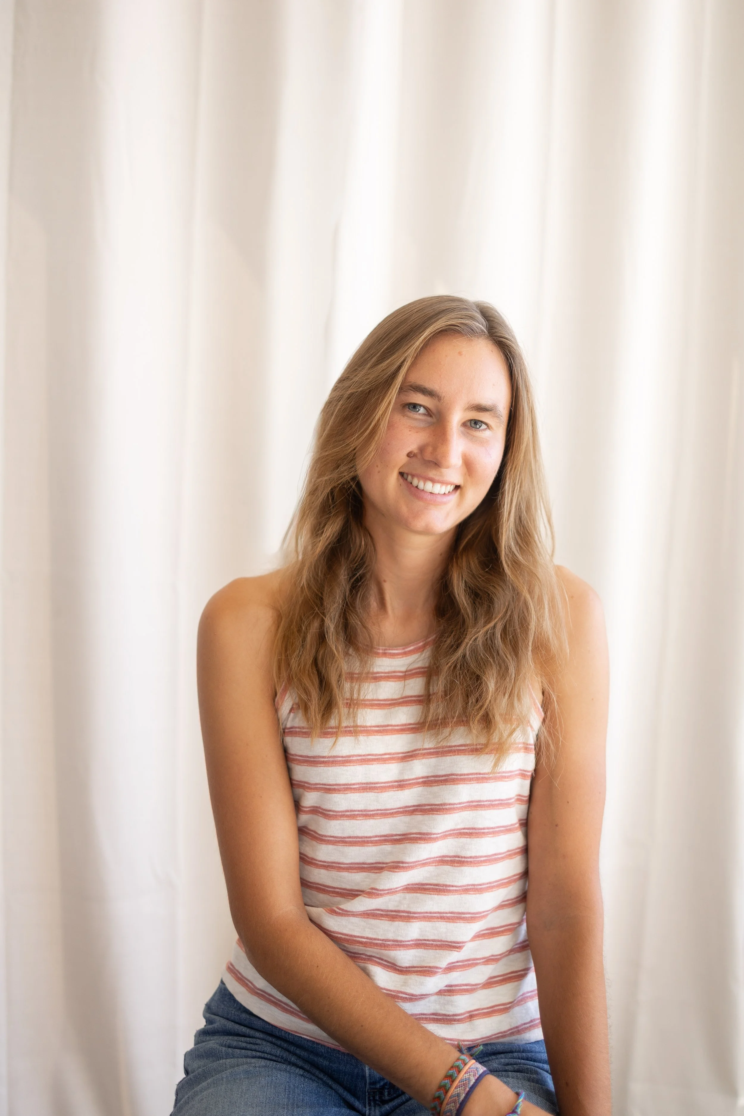 A young woman with long wavy brown hair smiling, wearing a striped sleeveless top and blue jeans, sitting in front of a white curtain.