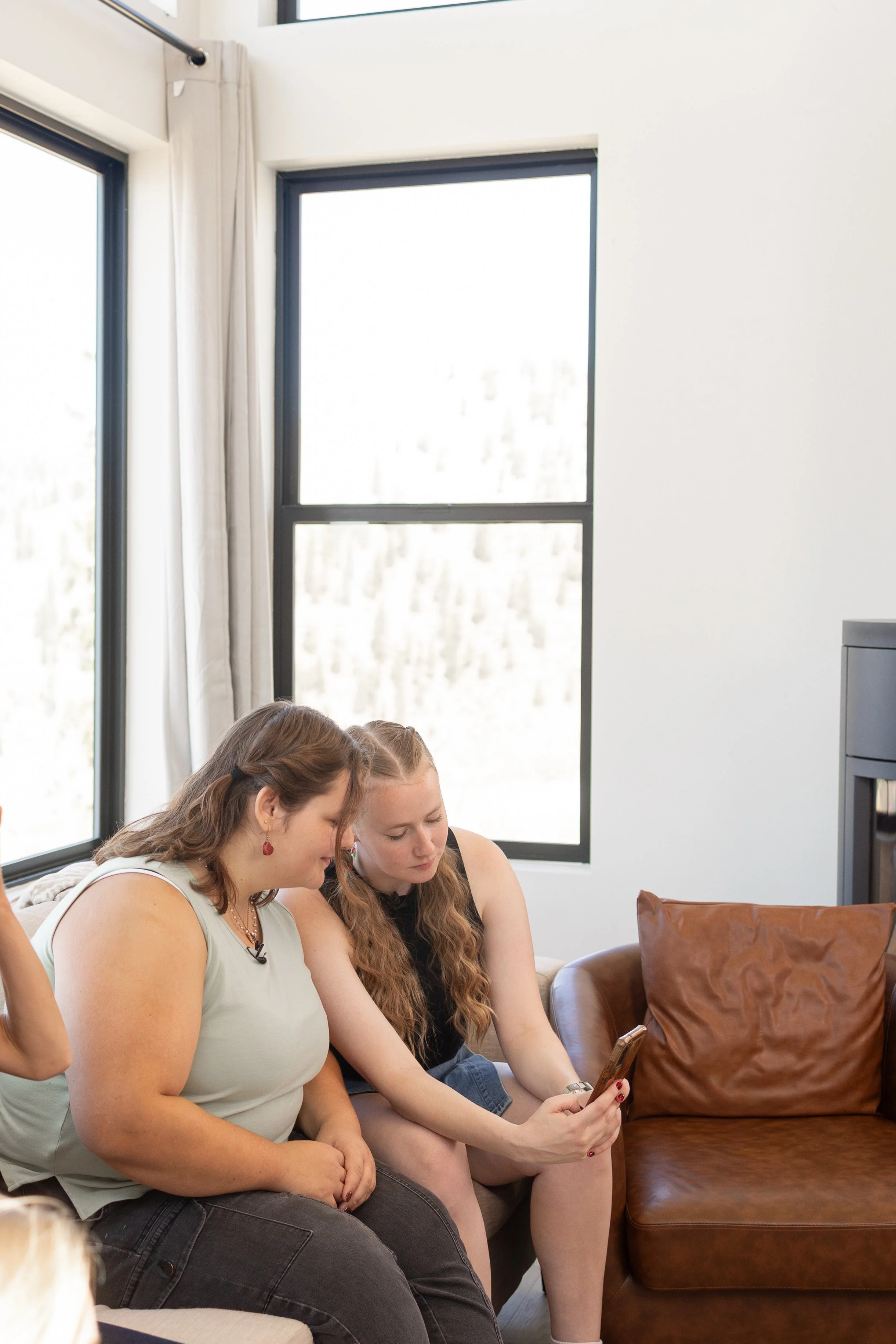 Two women sitting on a sofa, looking at a smartphone together in a brightly lit room with large windows and white walls.