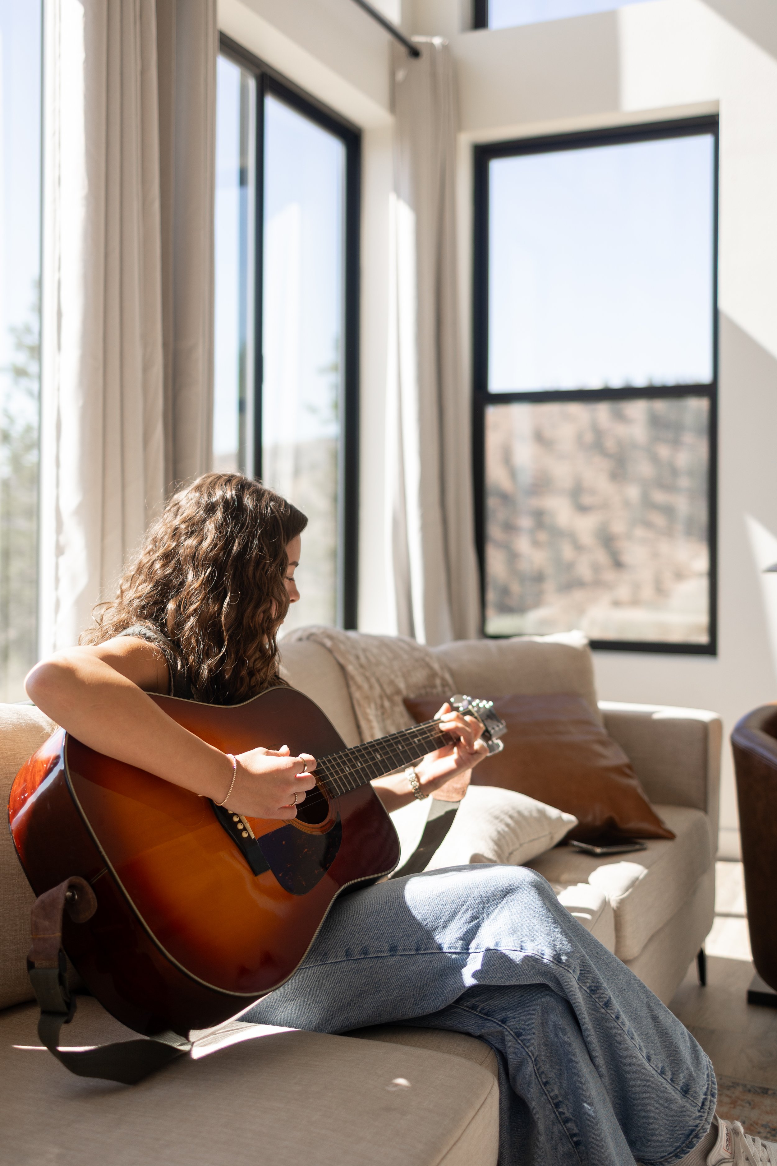 A woman with curly brown hair sitting on a beige sofa, playing an acoustic guitar, in a room with large windows and white curtains, sunlight illuminating the scene.