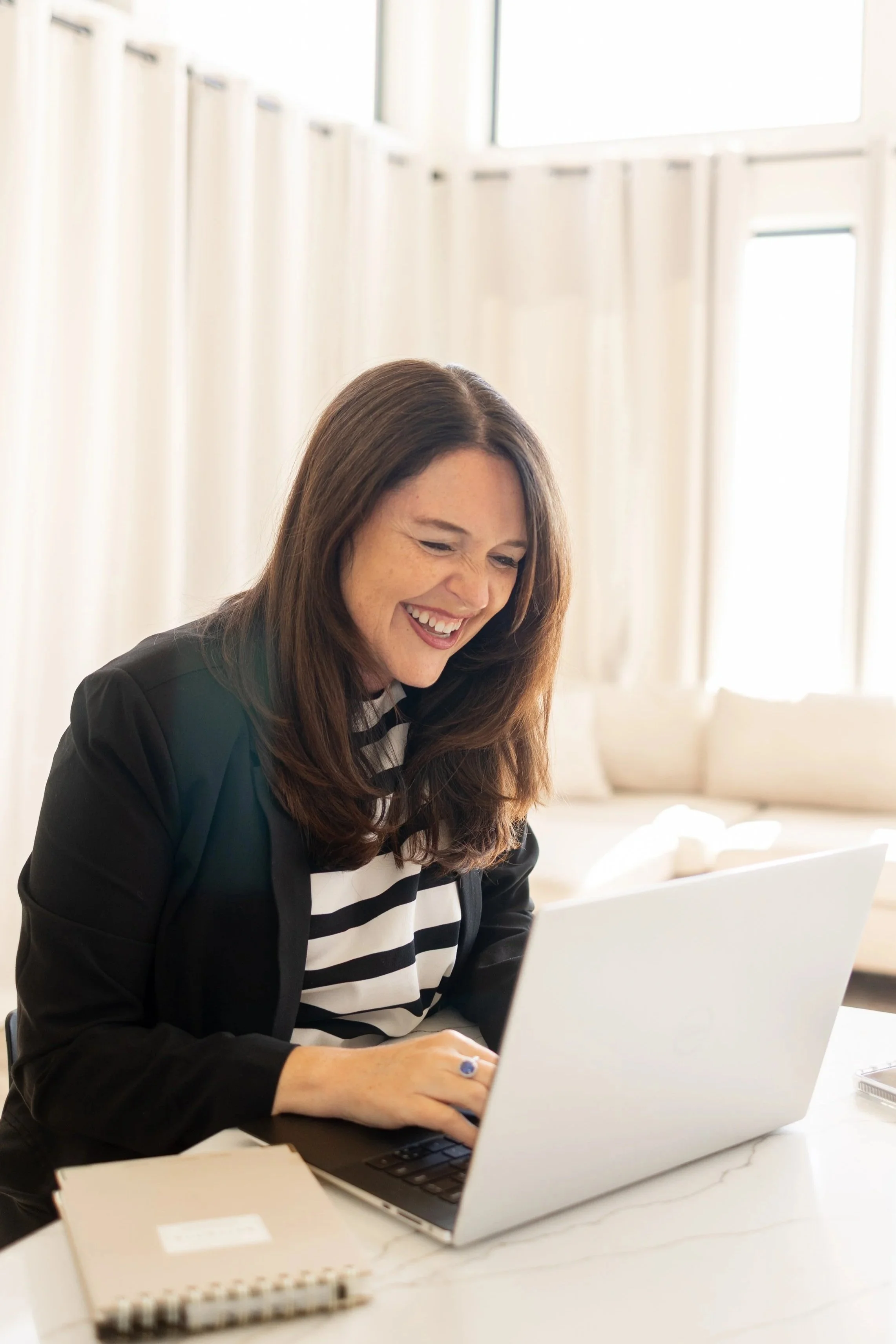 A woman with long brown hair smiling while working on a laptop at a white desk, with notebooks and papers nearby, in a bright room with white curtains and large windows.