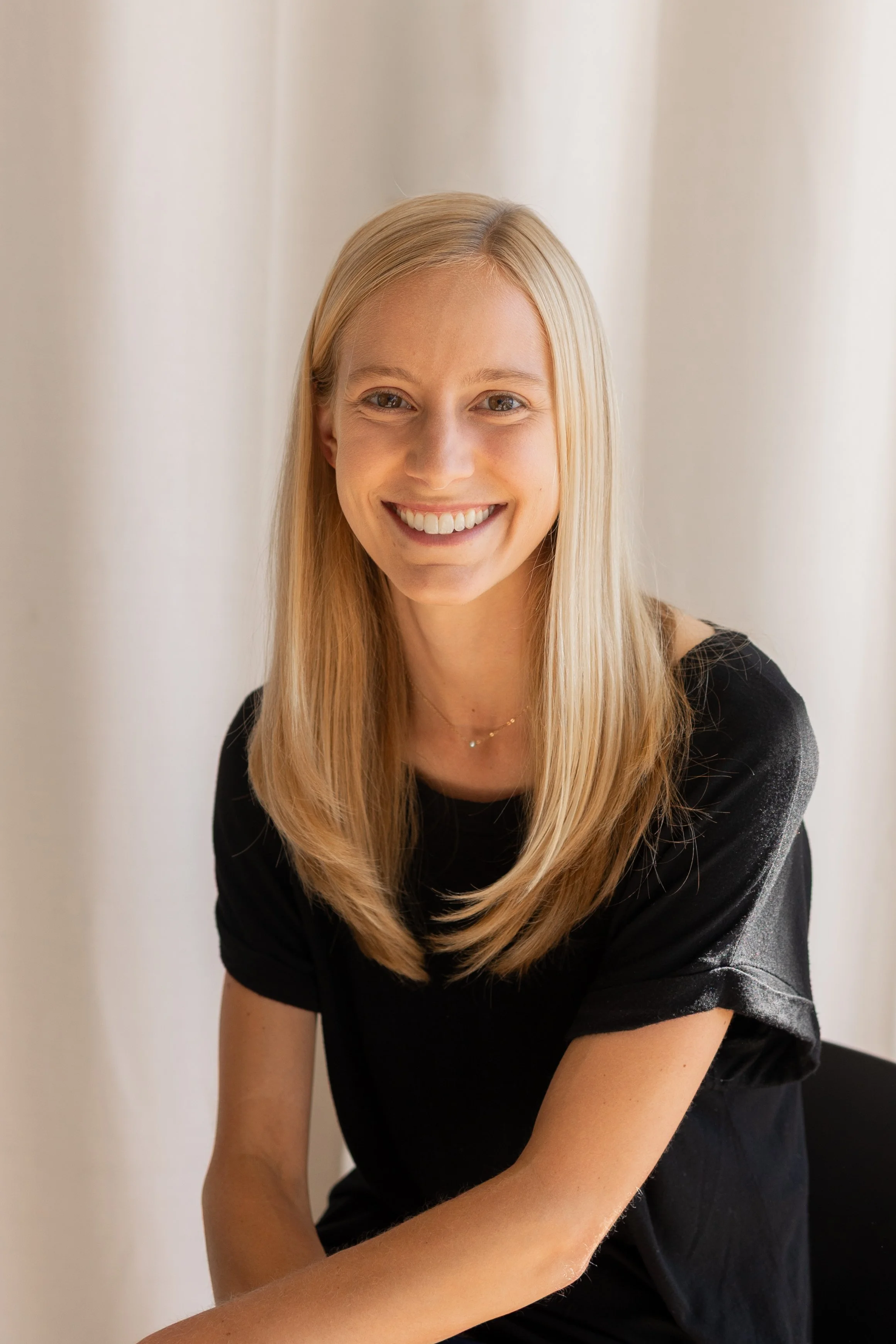A smiling young woman with long blonde hair and a black top, sitting against a white curtain background.