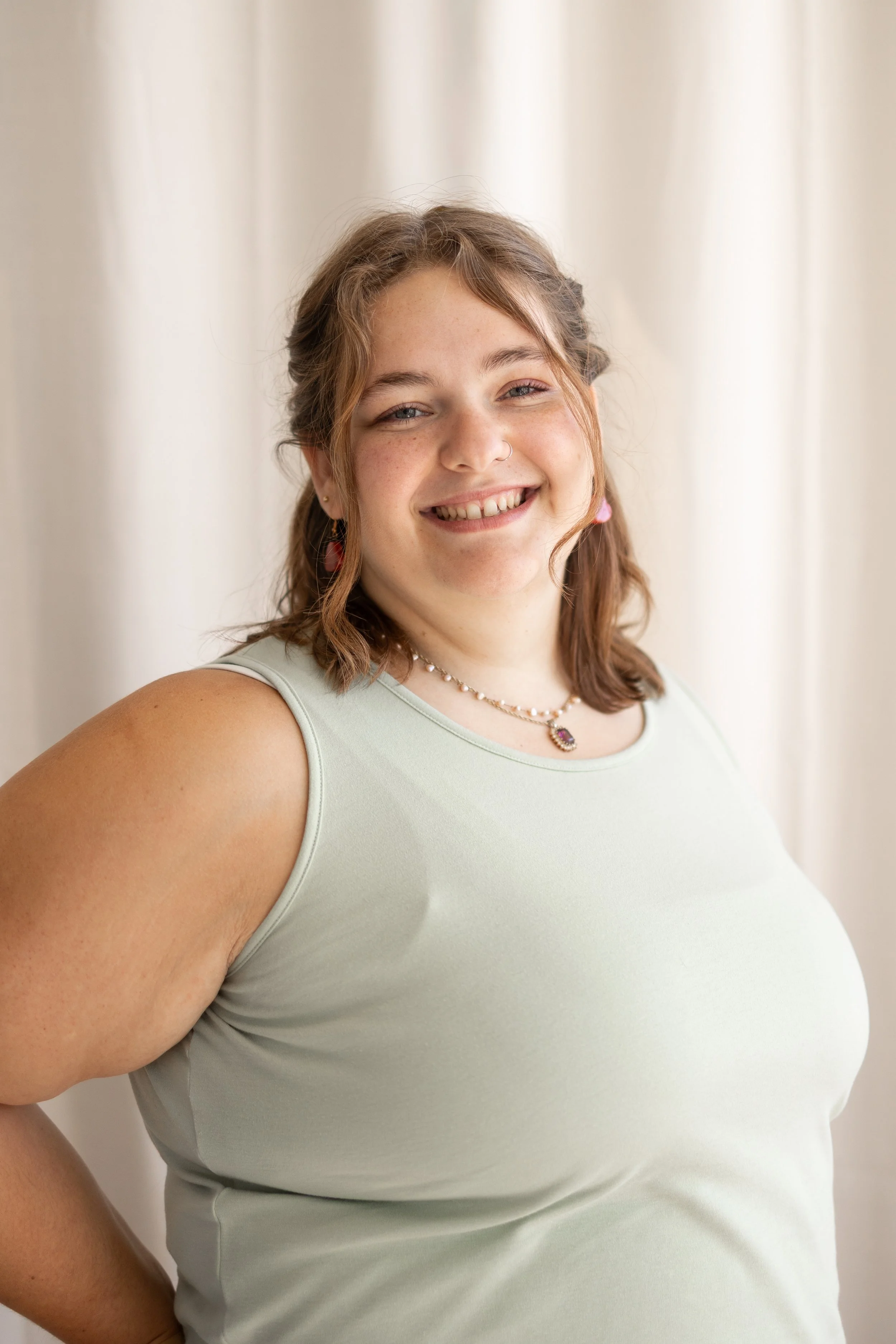 A woman with wavy brown hair, light skin, and blue eyes smiling while wearing a light green sleeveless top, layered necklaces, and a nose ring, standing against a neutral background.