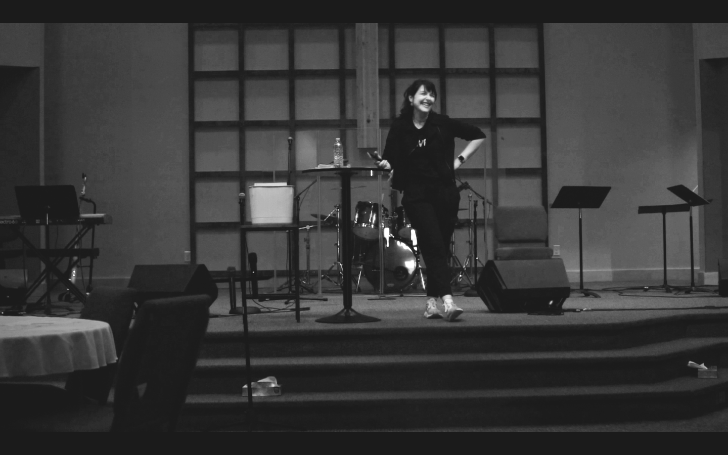 A woman standing on stage with musical equipment, smiling, holding a microphone, in a dimly lit setting.