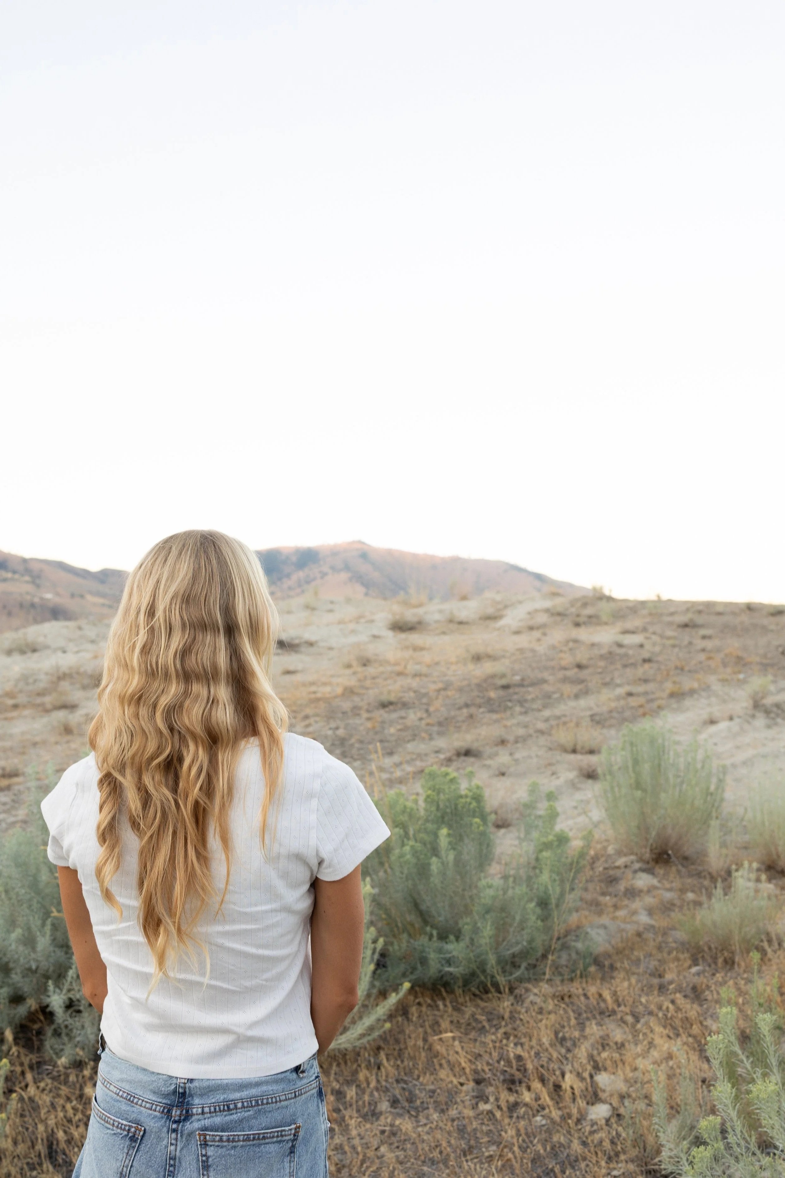 Back view of a woman with long wavy blonde hair standing outdoors in a desert landscape with hills and sparse vegetation, wearing a white T-shirt and denim jeans.