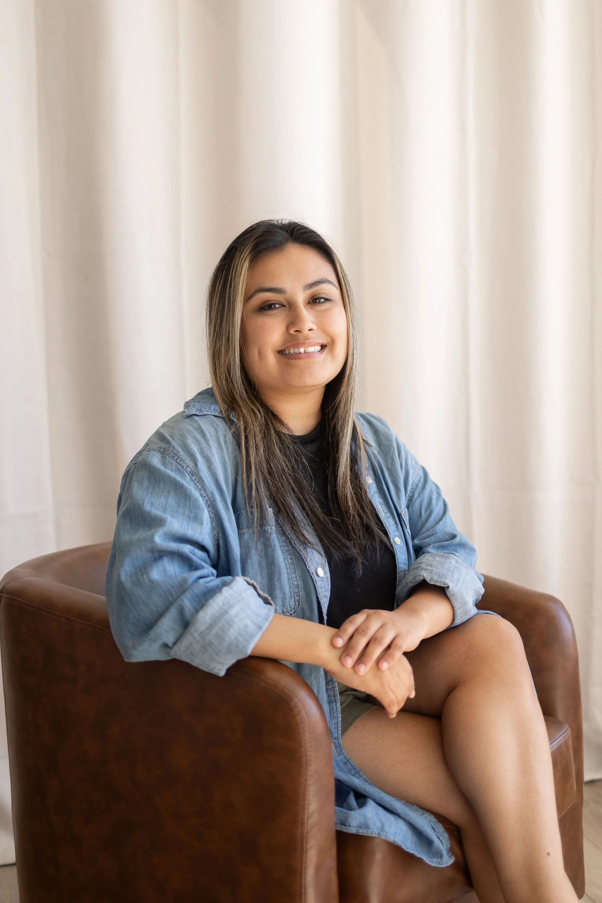 A young woman with long dark hair, wearing a denim jacket and shorts, sits on a brown leather chair in front of cream curtains, smiling and looking at the camera.