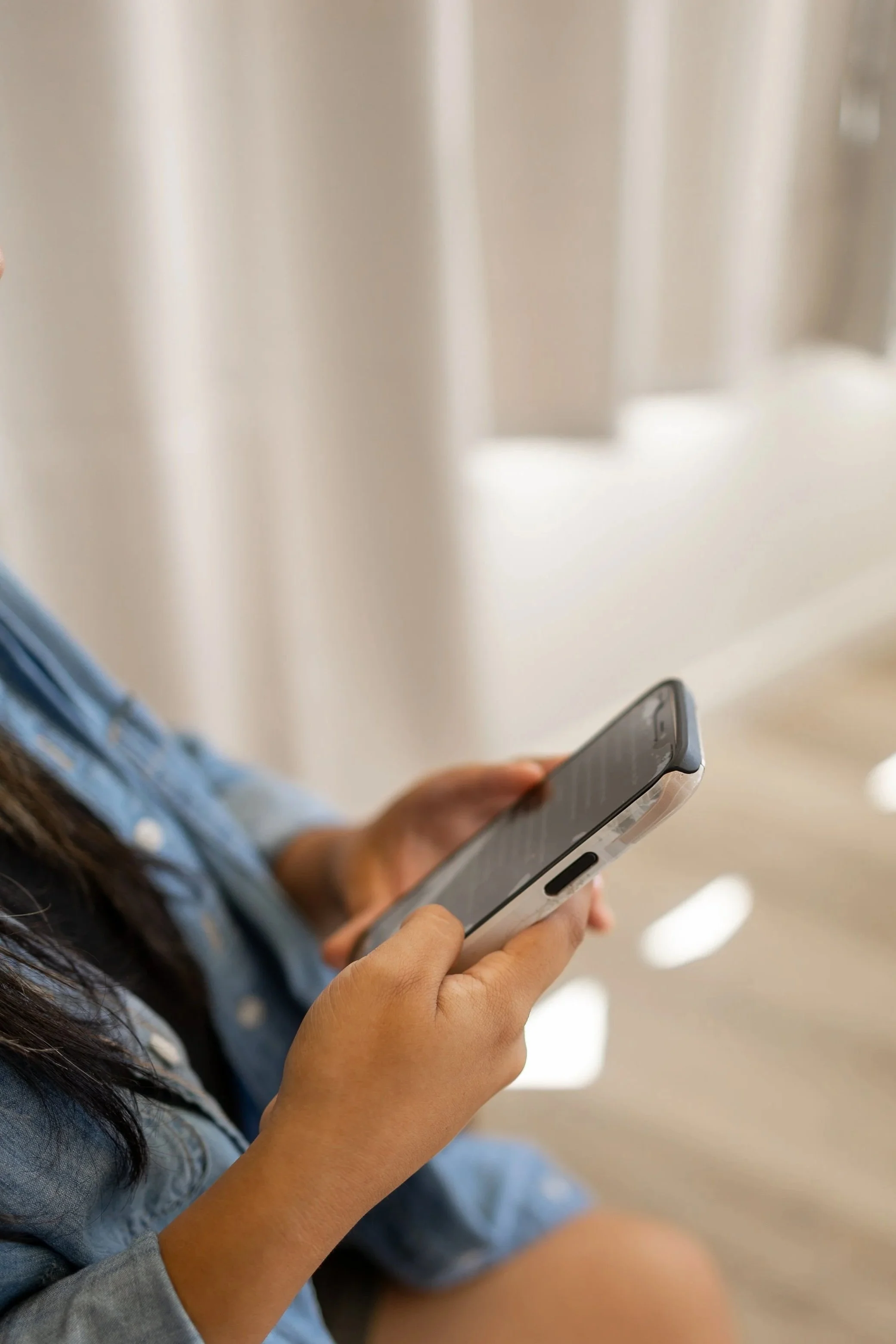 Close-up of a person holding a smartphone with both hands, wearing a gray long-sleeve shirt, with long dark hair visible. The background appears to be an indoor setting with a cream-colored wall.