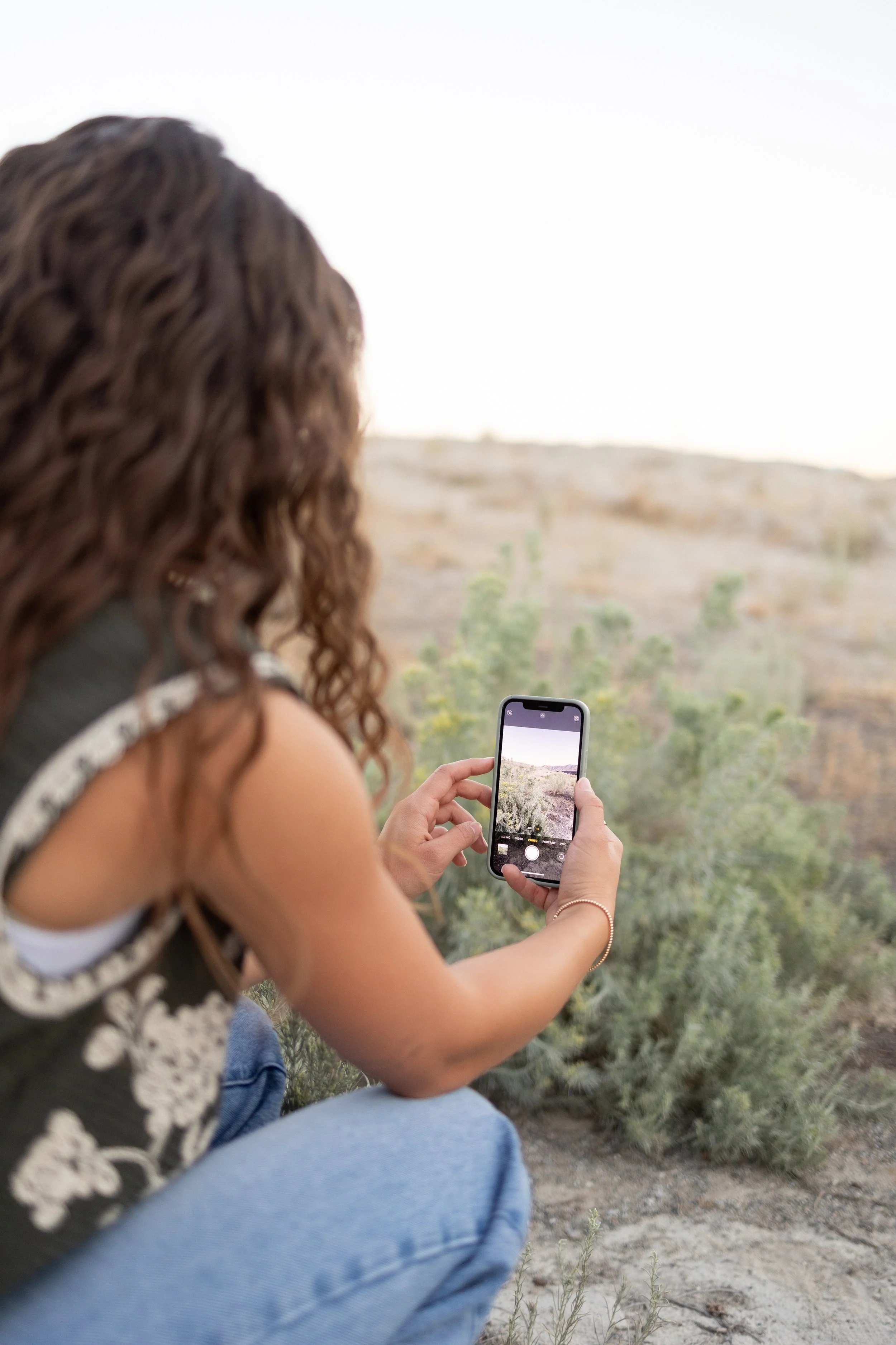 A woman with curly brown hair, wearing a black top with white embroidery, is sitting outdoors in a desert-like environment, taking a picture of the landscape with her smartphone. She is wearing blue jeans and a gold bracelet, and is surrounded by sparse green shrubs and rocky terrain.