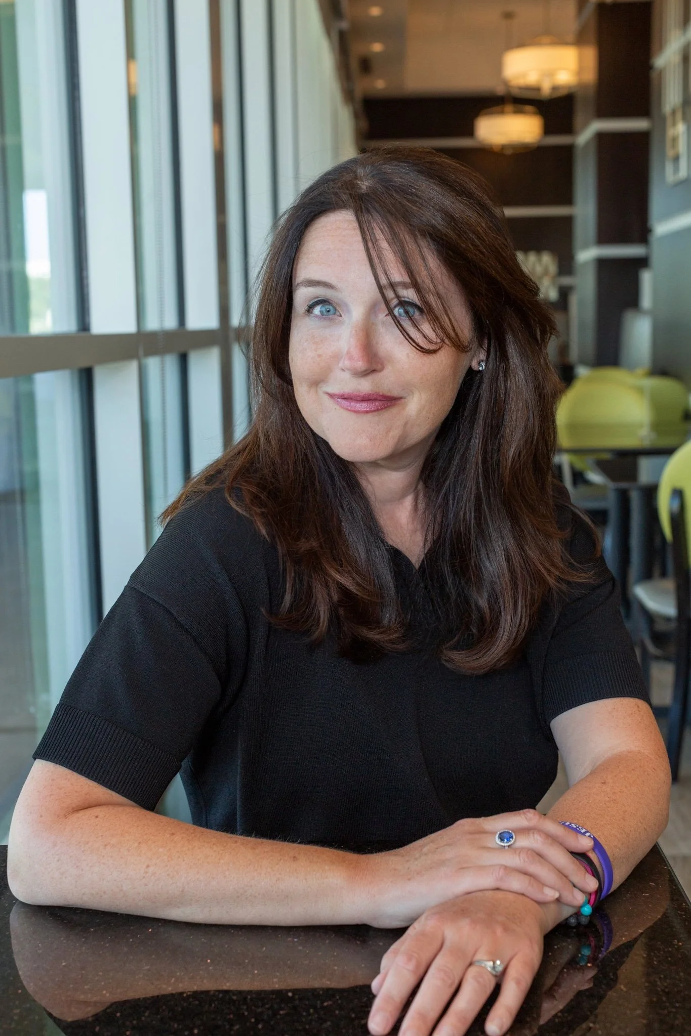 A woman with long brown hair and blue eyes sitting at a table in a modern indoor setting, wearing a black top, with colorful bracelets and a ring, looking at the camera.