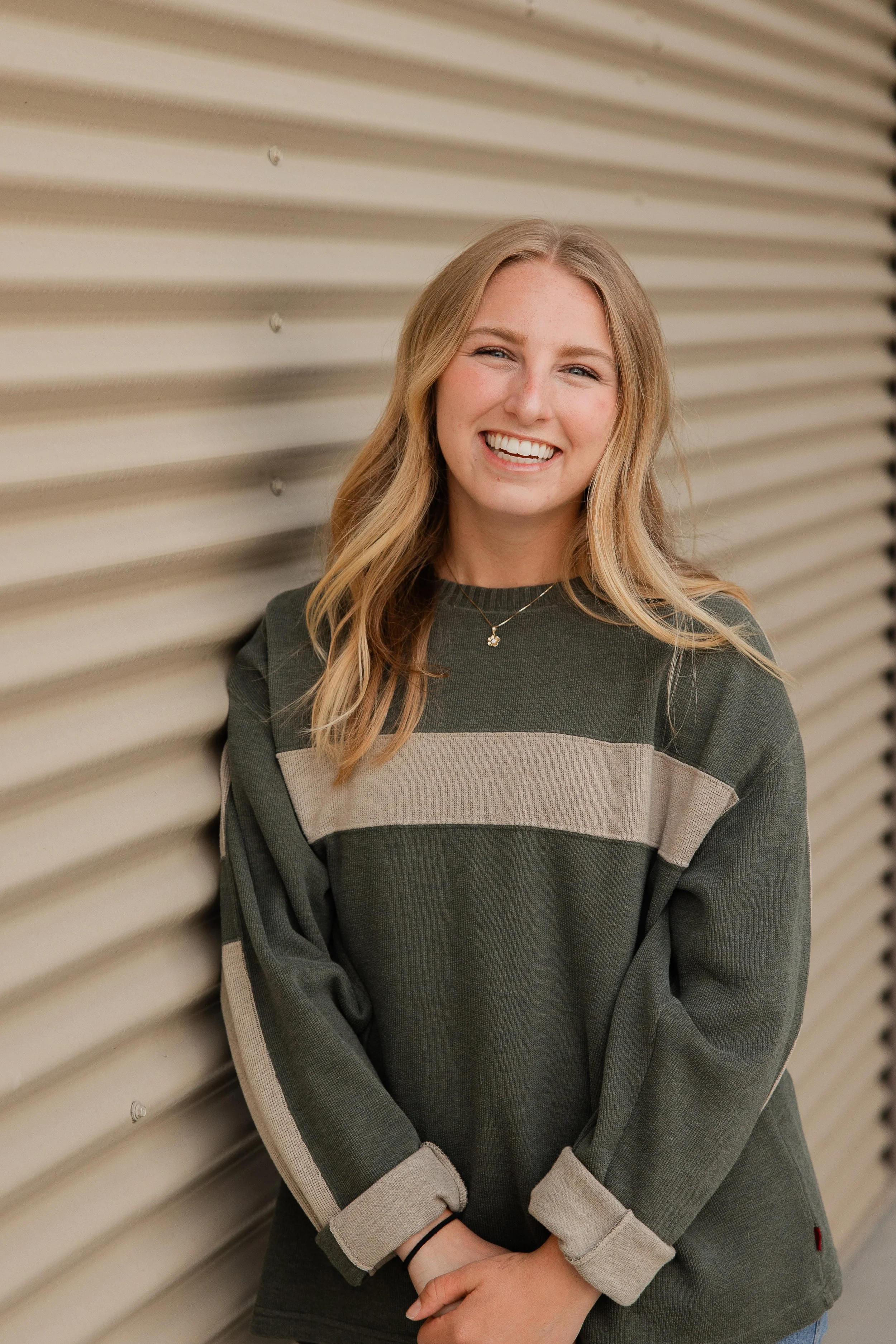 A young woman with long, wavy blonde hair smiling and looking at the camera while leaning against a beige, horizontal metal surface. She wears a dark green oversized sweatshirt with beige stripes and has her arms crossed with her hands clasped in front of her.