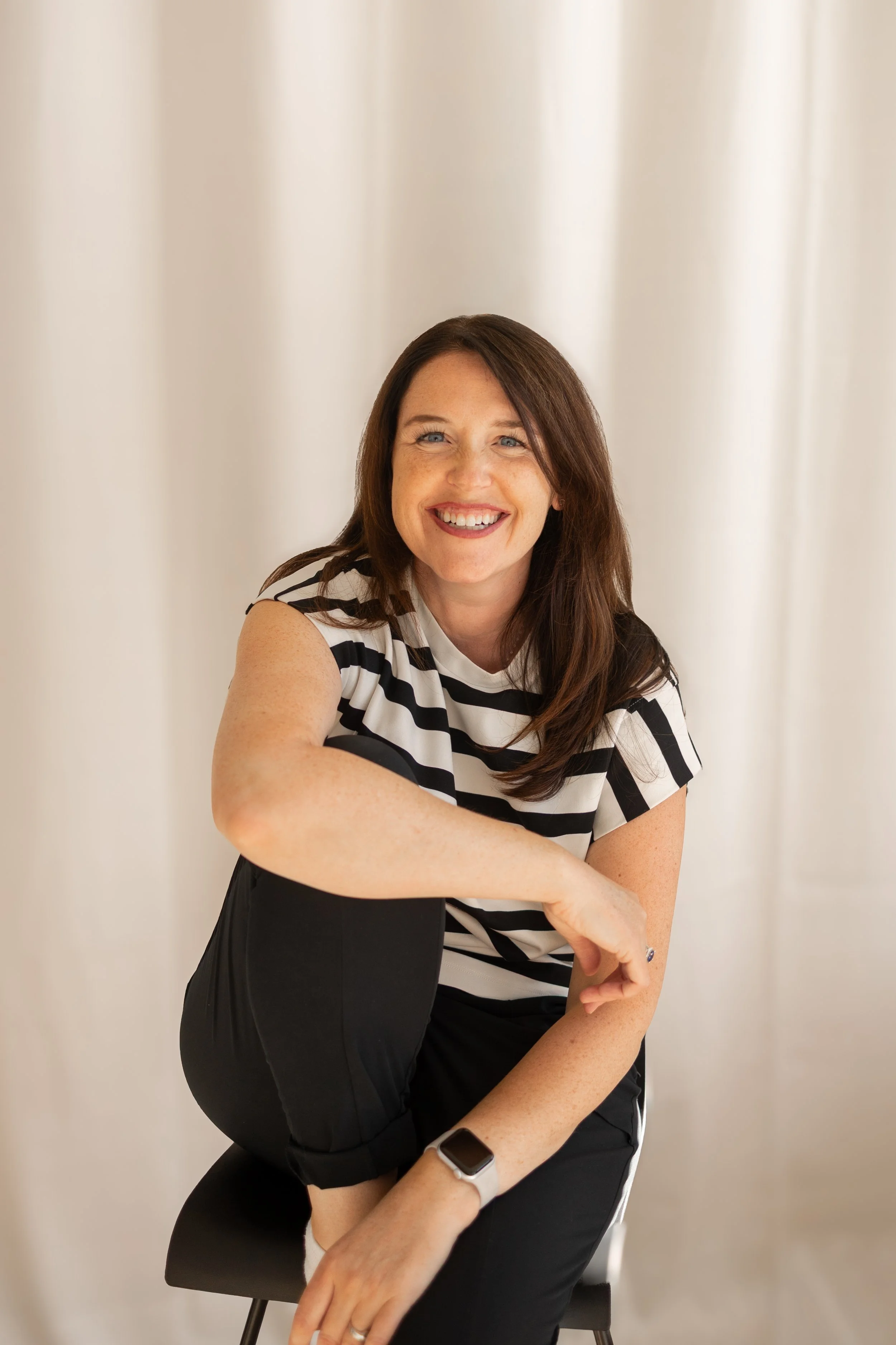 A woman with long brown hair, wearing a white and black striped short-sleeve shirt and black pants, smiling and sitting on a chair with a plain white curtain background.