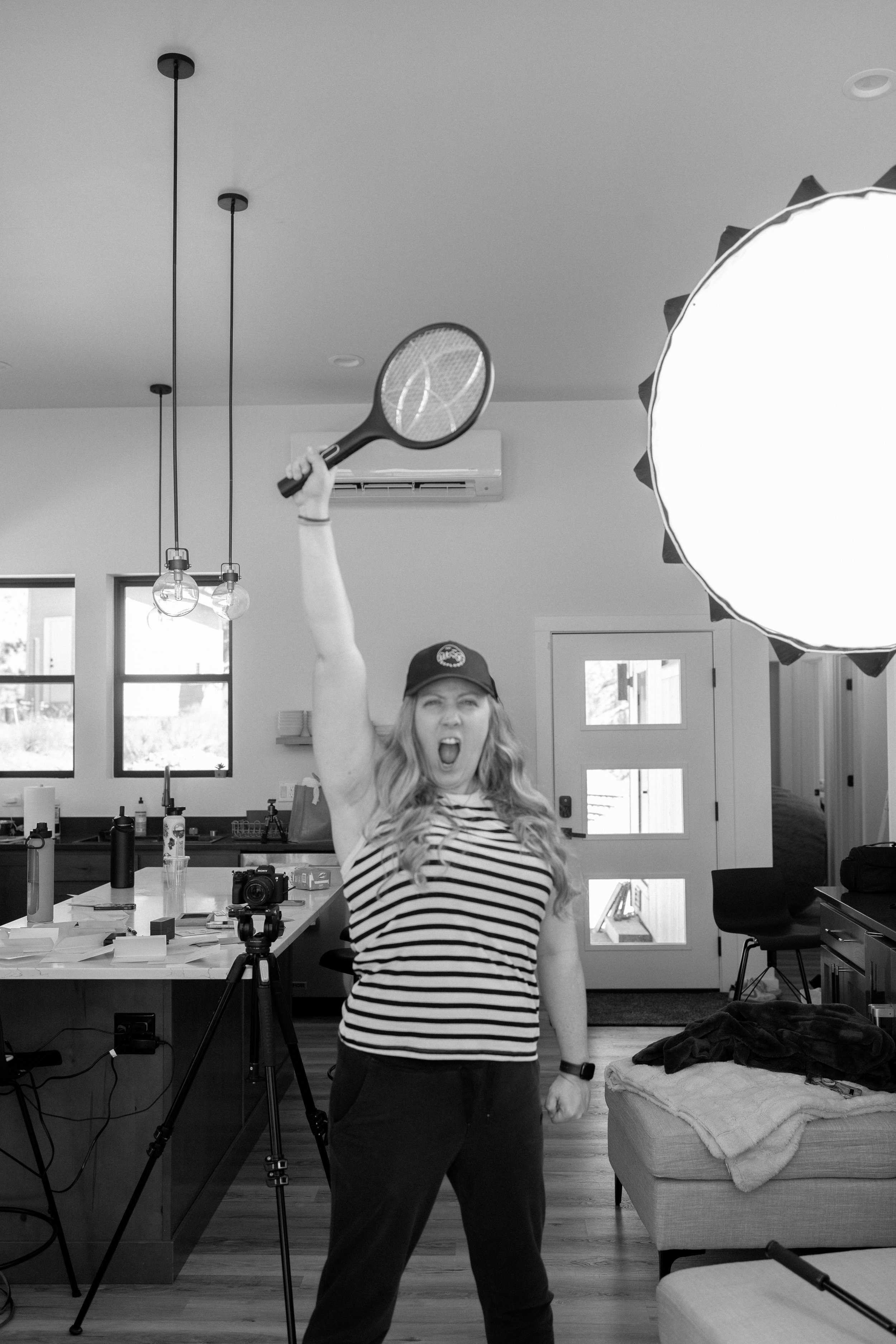Woman in striped shirt holding tennis racket raised above her head, standing in a kitchen with cameras, a tripod, and photography equipment around her, a softbox light to the right, and a ceiling fan and air conditioner in the background.
