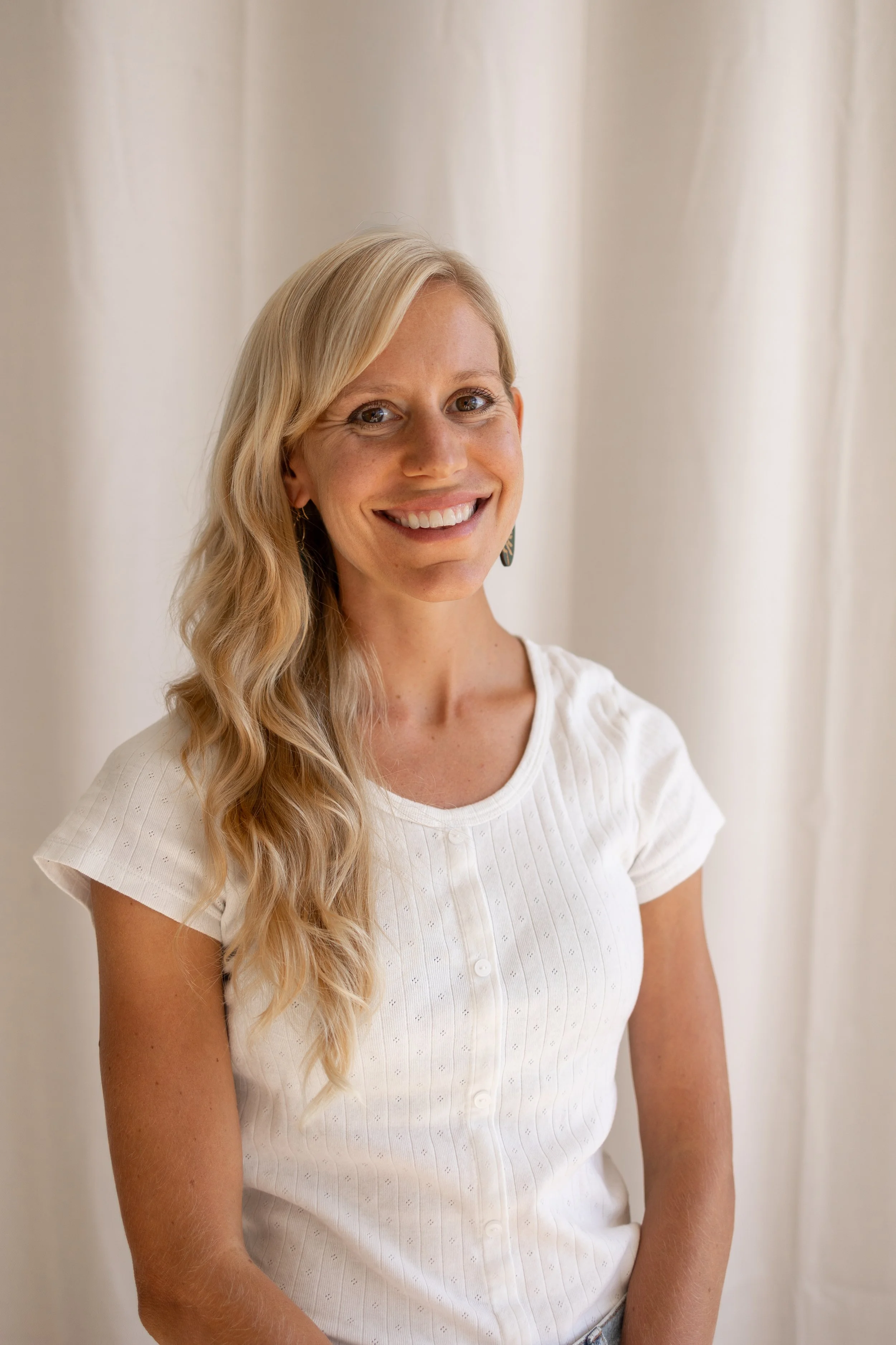 A woman with long, wavy blonde hair smiling, wearing a white short-sleeved shirt with buttons, standing in front of a neutral background.