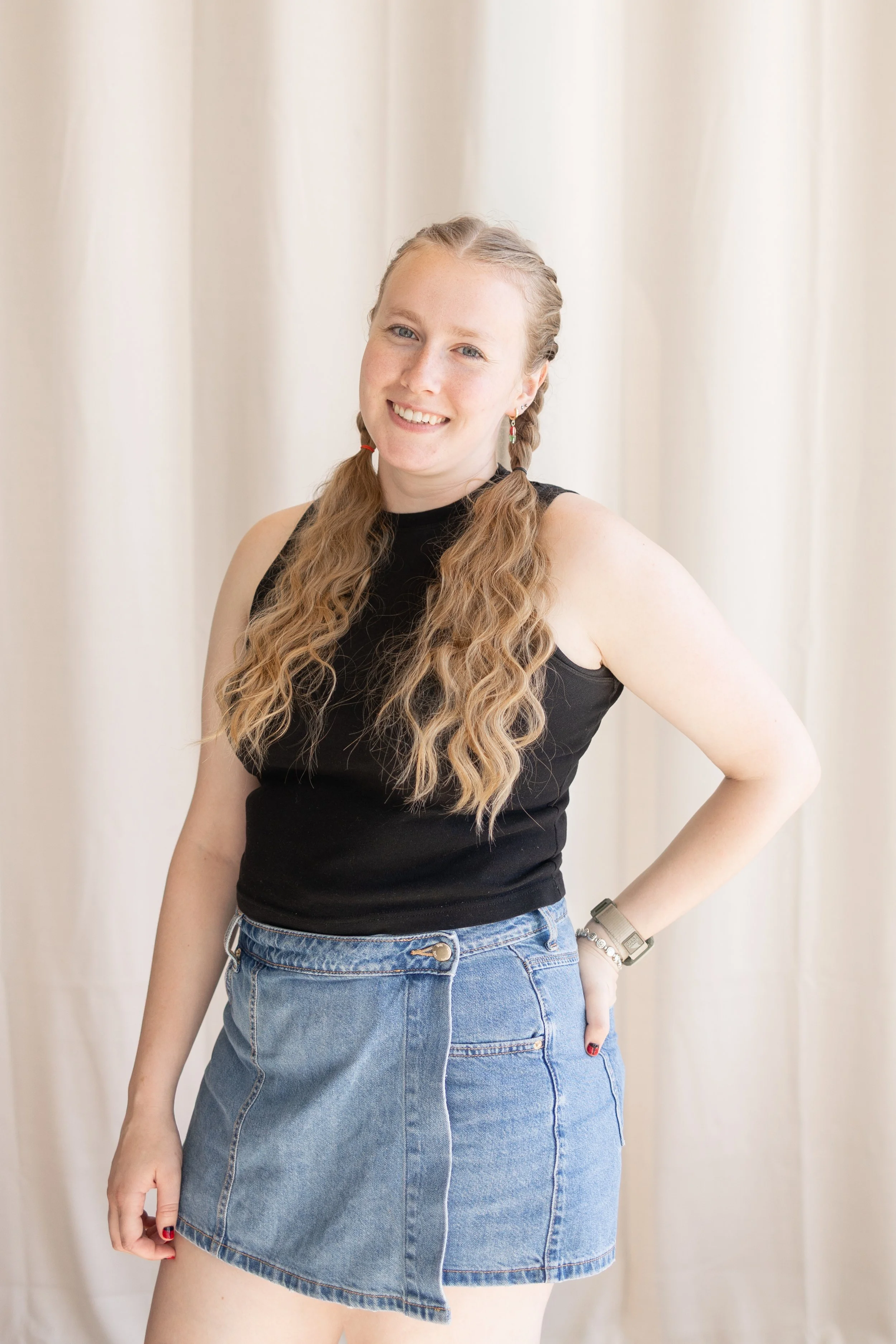 A young woman with long wavy blonde hair styled in braids, wearing a black sleeveless top and high-waisted denim skirt, standing with her hand on her hip in front of cream-colored curtains.