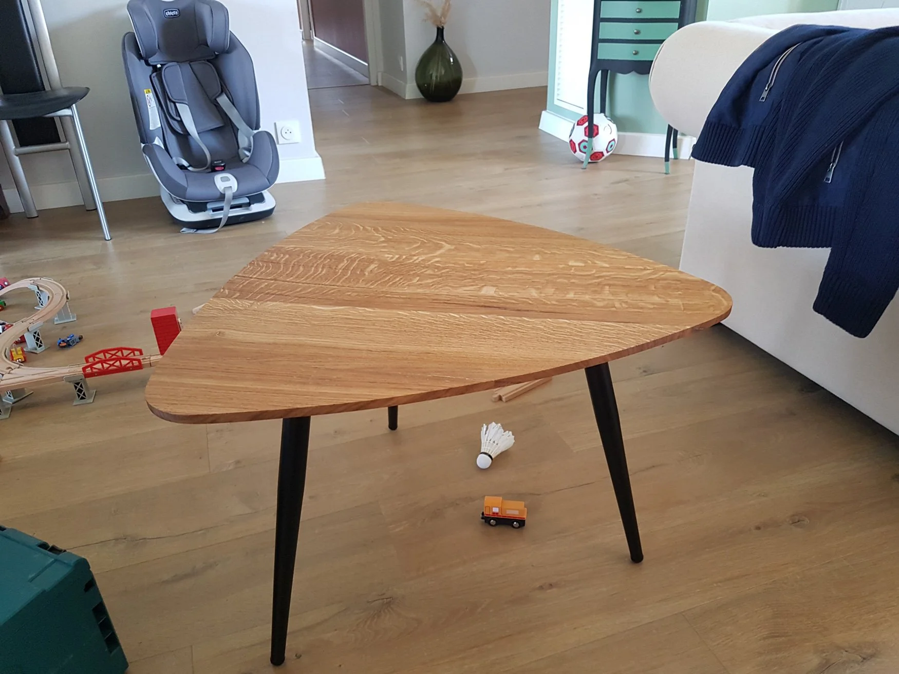 Intérieur d'une pièce avec une table en bois, un fauteuil d'enfant gris, un arbre de basketball, des jouets de train en dessous, et une plante verte dans un vase à l'arrière.