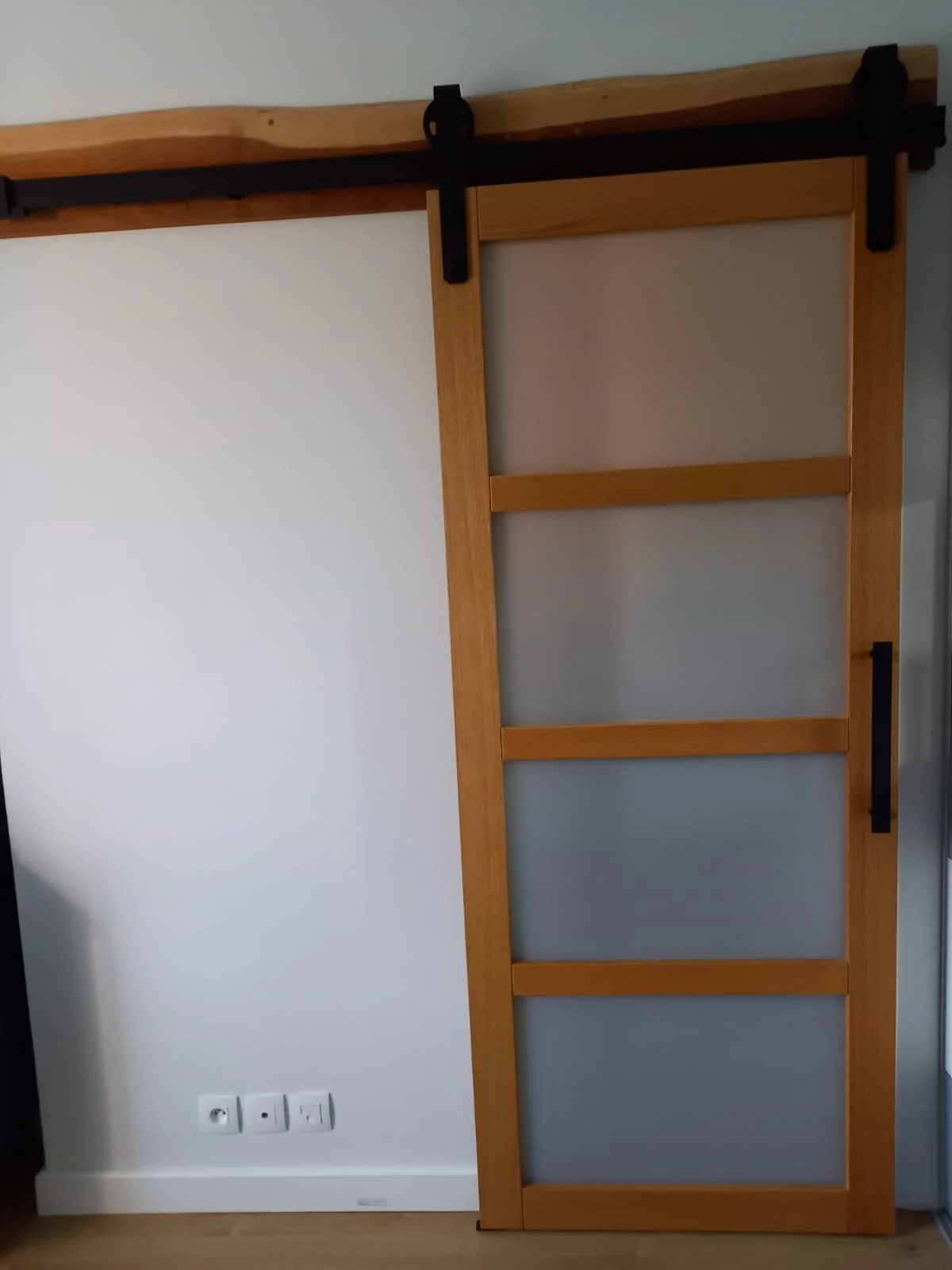 Une porte de style coulissante en bois avec verre, montée sur un rail noir, dans une pièce avec un mur blanc et une prise électrique.